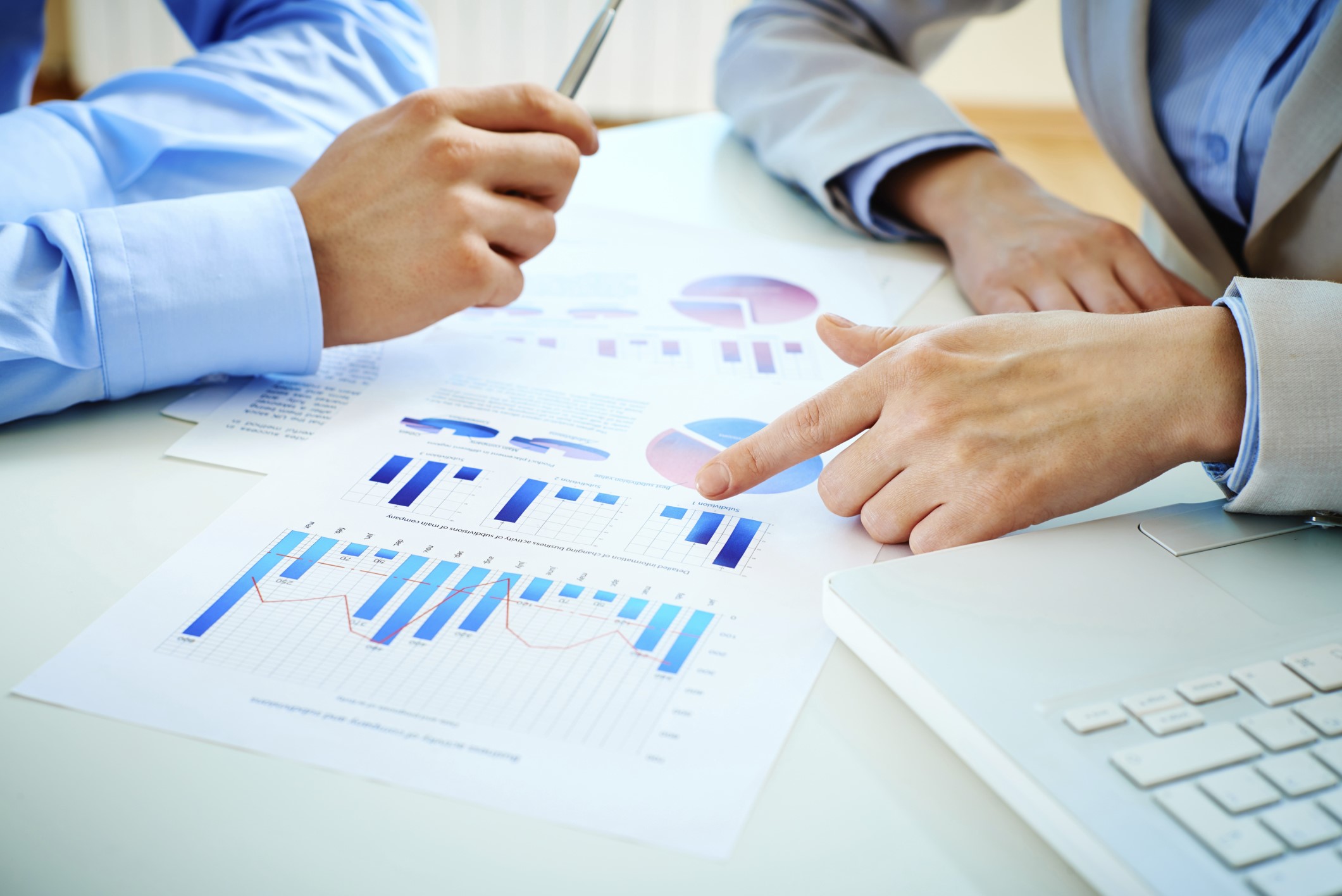 Two people analyzing printed charts and graphs on a table, with one person pointing at a pie chart and the other holding a pen, with a laptop nearby.
