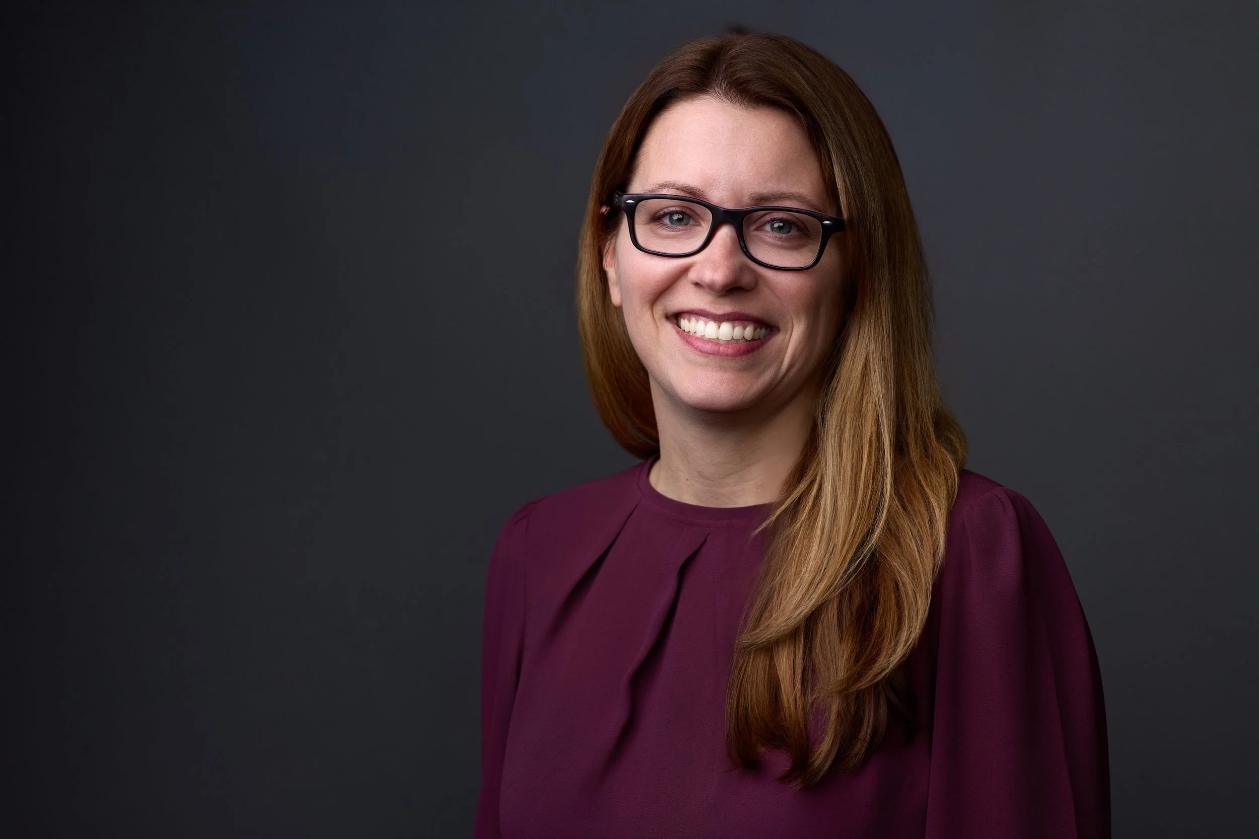A woman with long brown hair wearing black glasses and a purple blouse, smiling against a dark gray background.