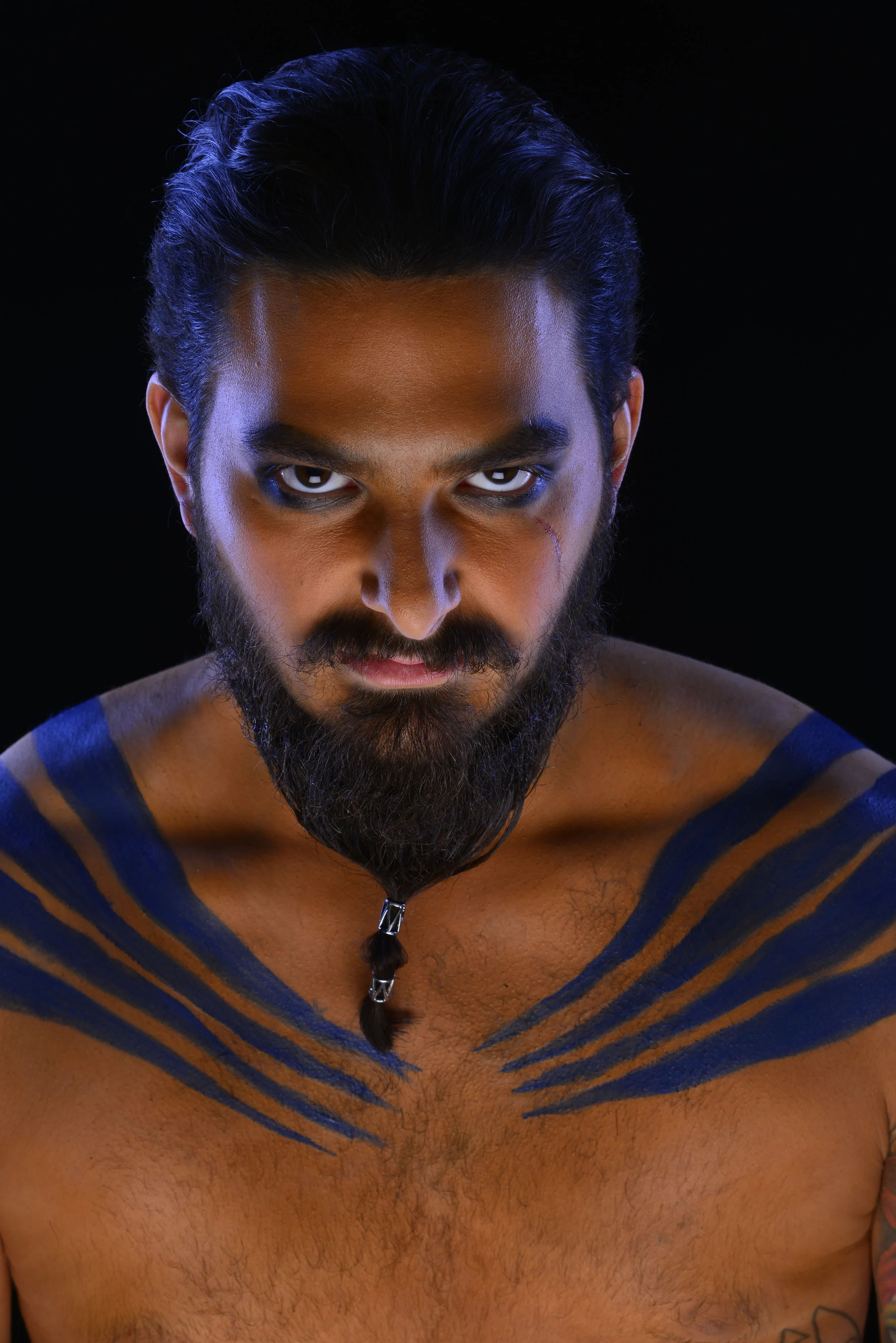 A man with a serious expression, dark eyes, a beard, and long dark hair tied with a small braid, posing shirtless with blue face paint on his shoulders and chest against a dark background.