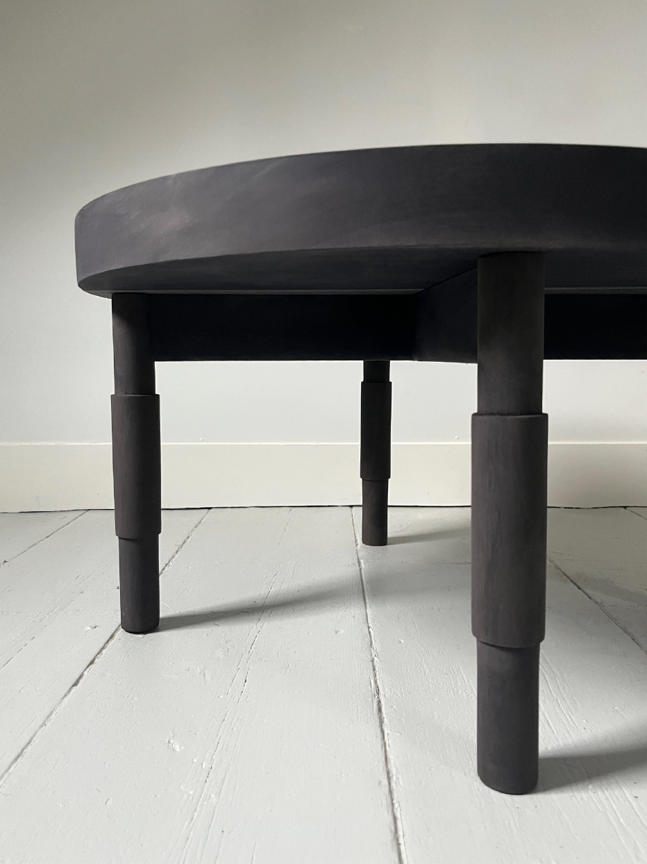 Close-up view of a black wooden table with round edges and four thick cylindrical legs, positioned on a white wooden floor against a plain white wall.