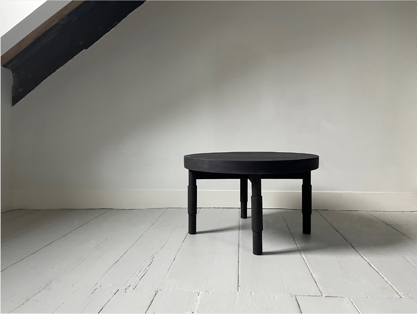 Small black round table with three legs in a minimal room with white wooden floor and white wall, no other furniture or decorations.