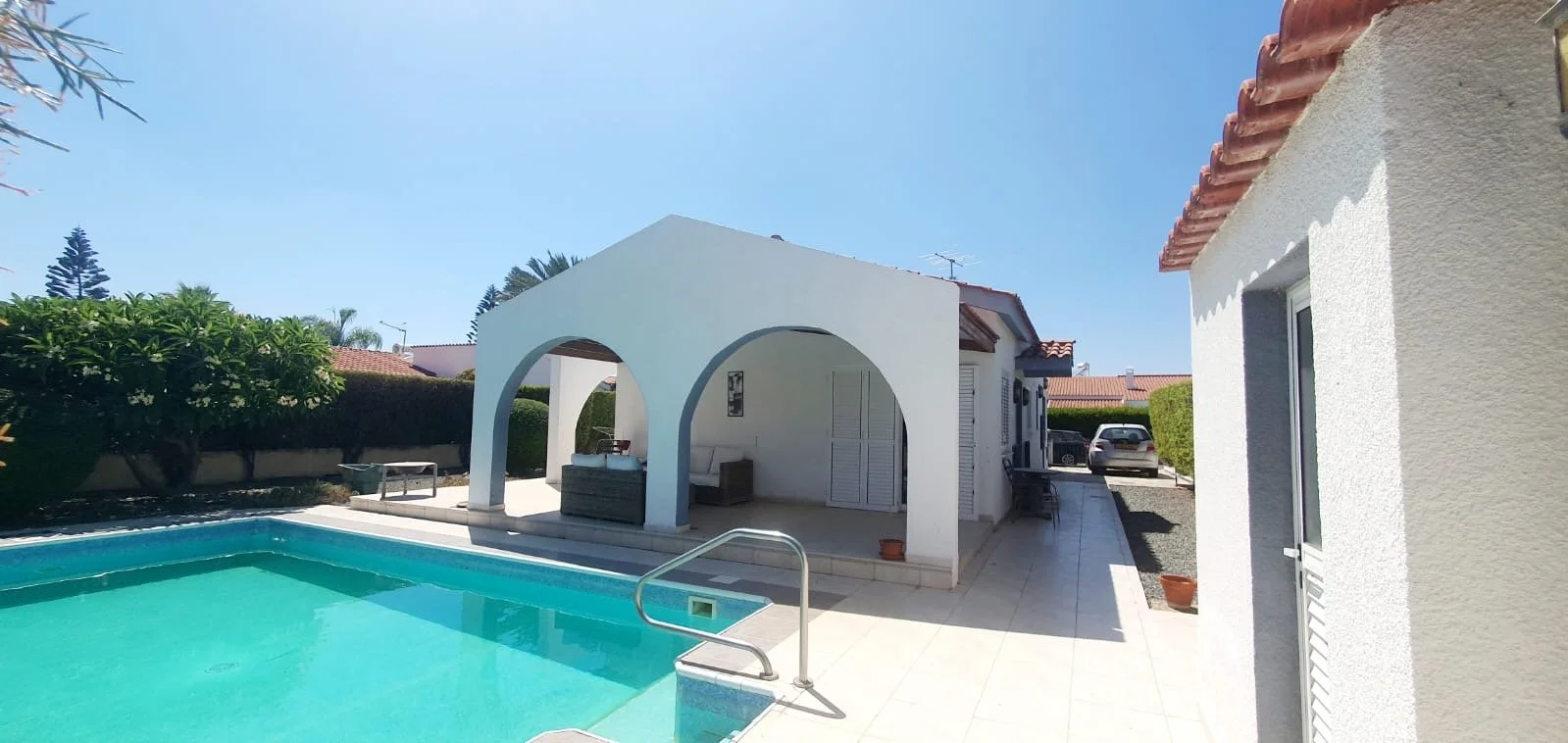 Outdoor view of a backyard with a swimming pool, white houses, and a seating area under an arched structure, with clear blue sky.