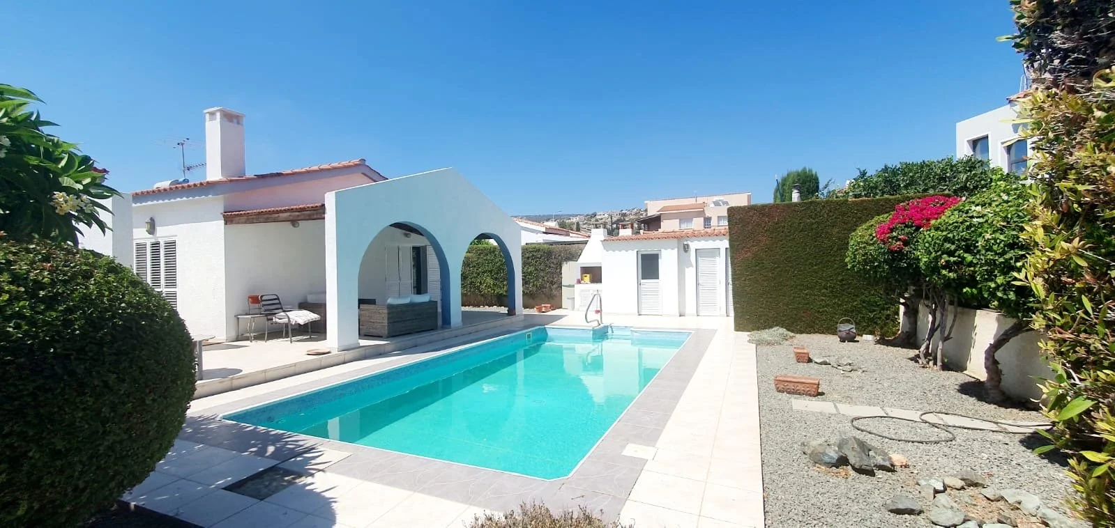 A backyard with a swimming pool, surrounded by bushes, trees, and a white house with a red-tiled roof.