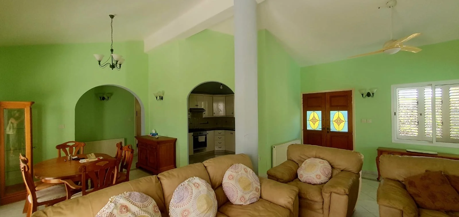 Living room with green walls, beige sofas with circular patterned cushions, a dining table with chairs, a chandelier, wooden cabinet, and a view of the kitchen through arched doorways.