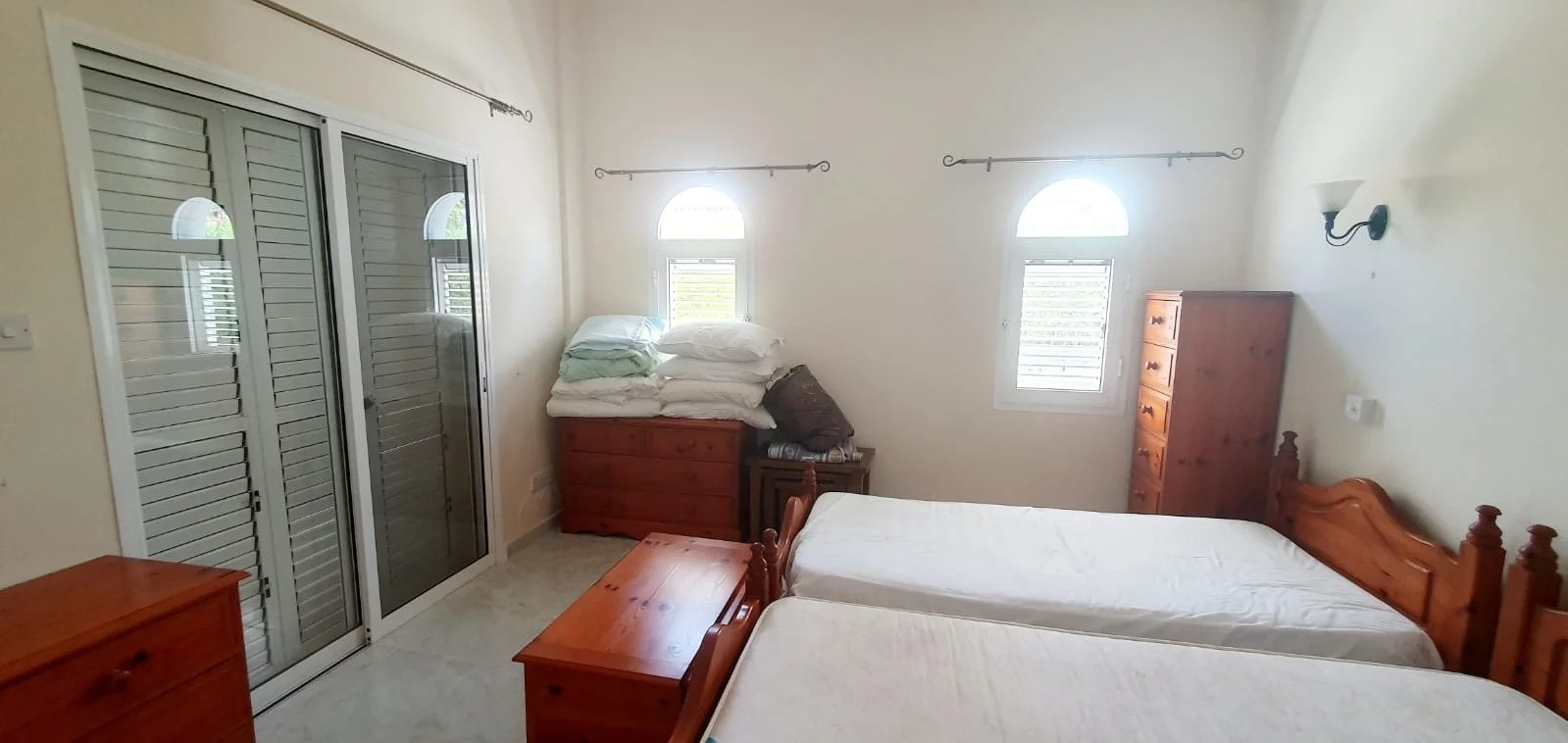 A bedroom with two single beds, a wooden dresser, a tall wooden wardrobe, and two windows with shutters. There are some folded bedding and pillows on a chest at the end of one bed. The room has a light-colored wall and tiled floor.
