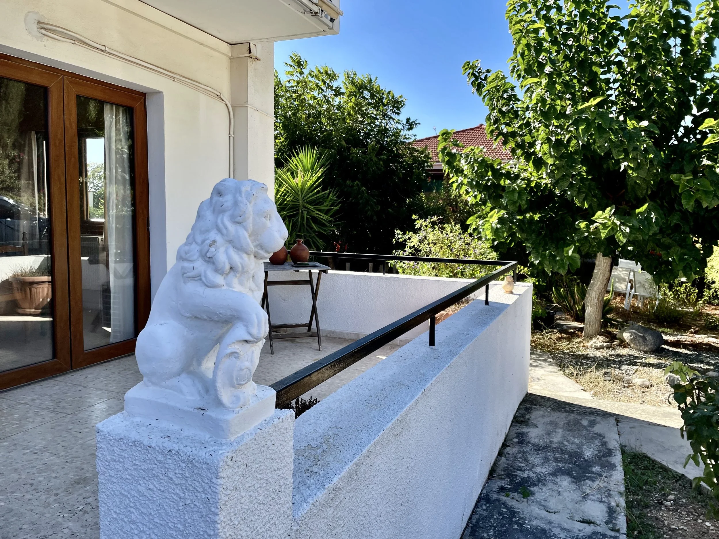 White stone lion statue on a small pedestal on a balcony with a black railing, next to a sliding glass door, with greenery and trees in the background and a clear blue sky.