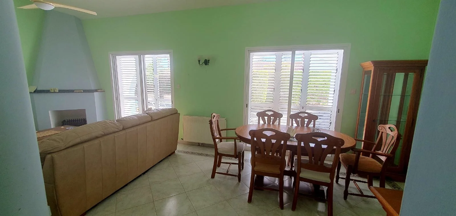 Living room with green walls, a beige sofa, a wooden dining table with six chairs, a glass-fronted wooden cabinet, windows with white shutters, and a tiled floor.