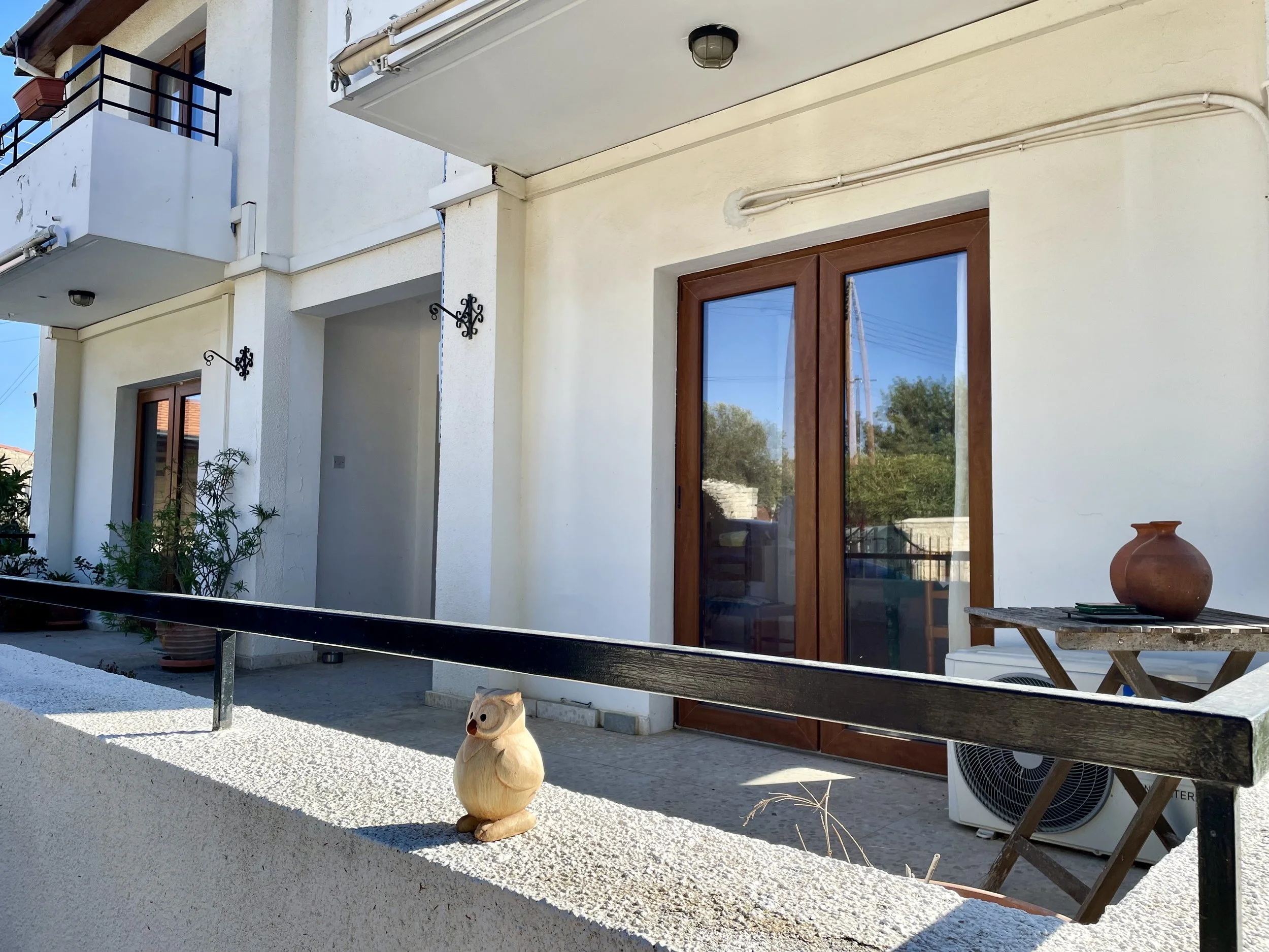 View of a balcony with a wooden door, a small table with a clay pot, garden plants, and a decorative owl figurine on the wall, with reflections of trees and power lines in the glass door.