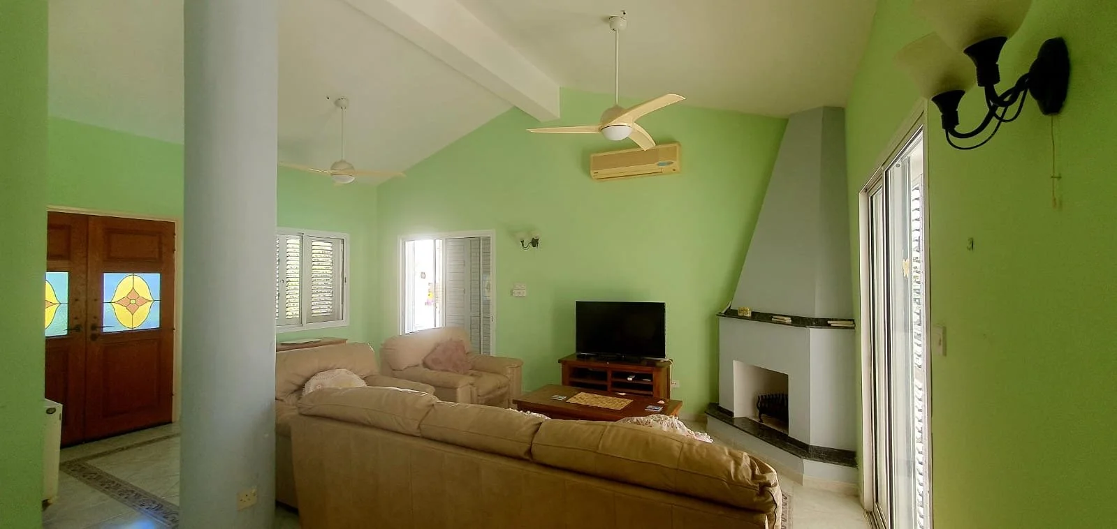 Living room with light green walls, beige sofas, a wooden TV stand with a flat-screen TV, a white fireplace, windows, and sliding glass doors.