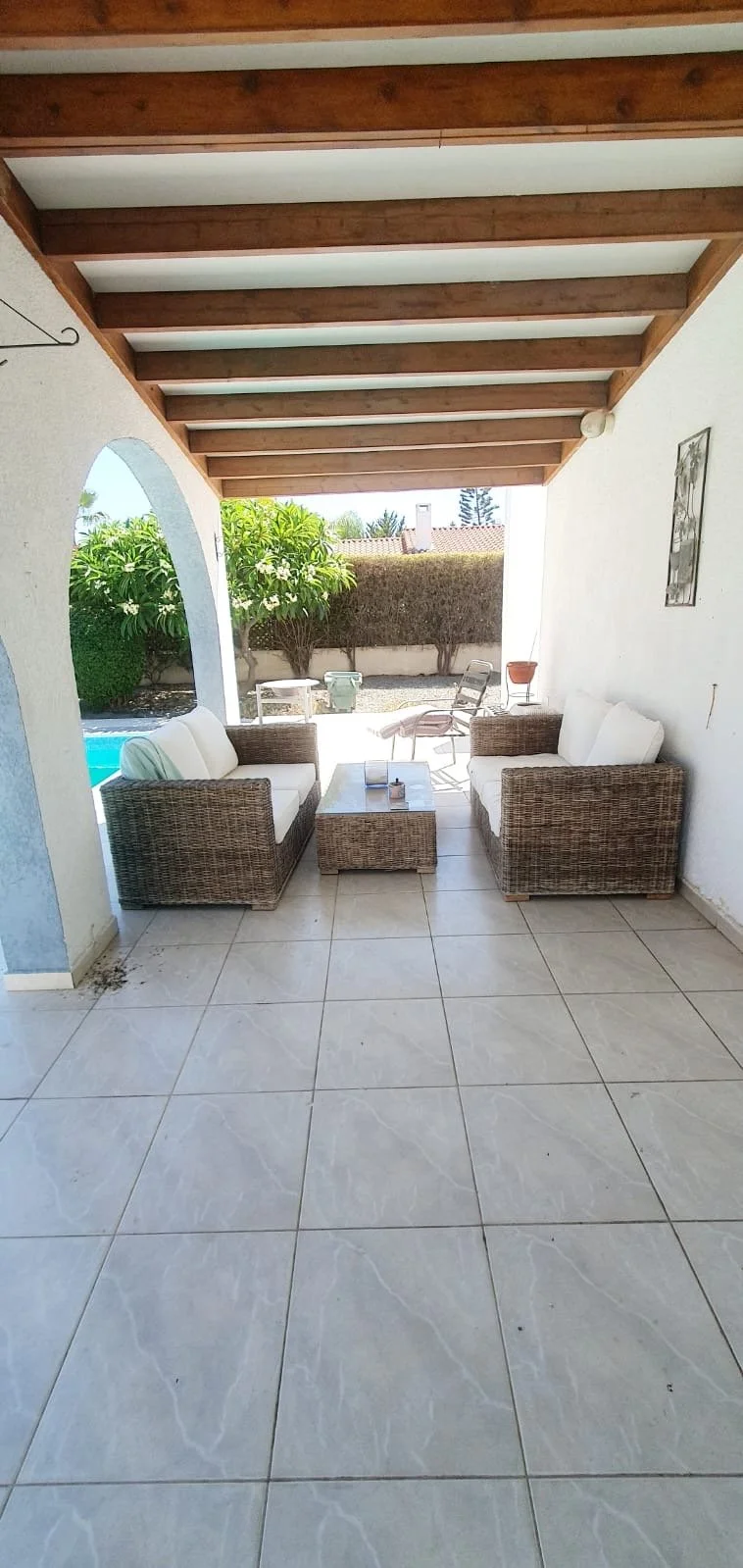 Patio area with wicker sofas and a coffee table under a wooden pergola, with a backyard and pool visible in the background.