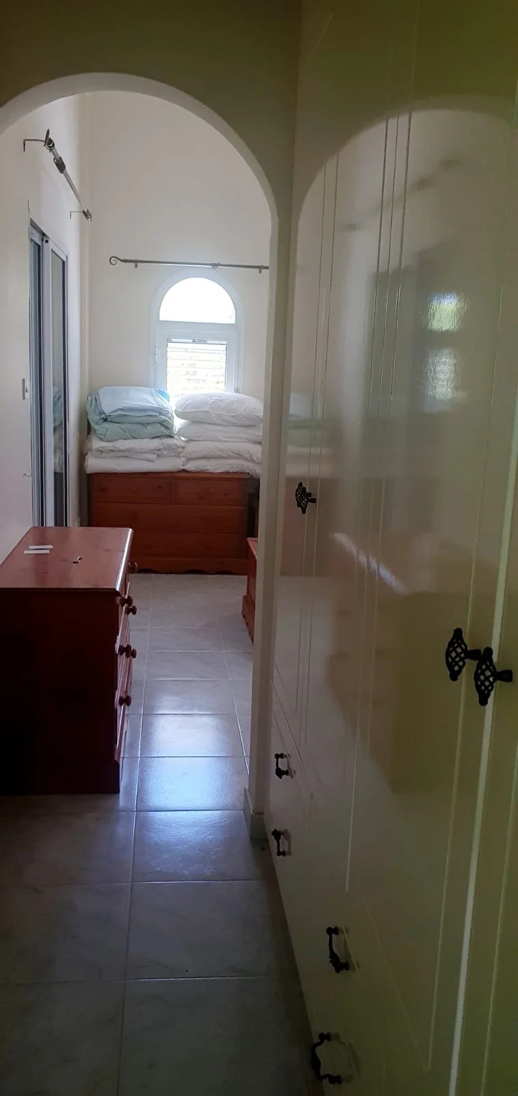 A small bedroom with a stack of pillows and bedding on a wooden bed frame, a window with sunlight, a dresser, and tall closet cabinets.