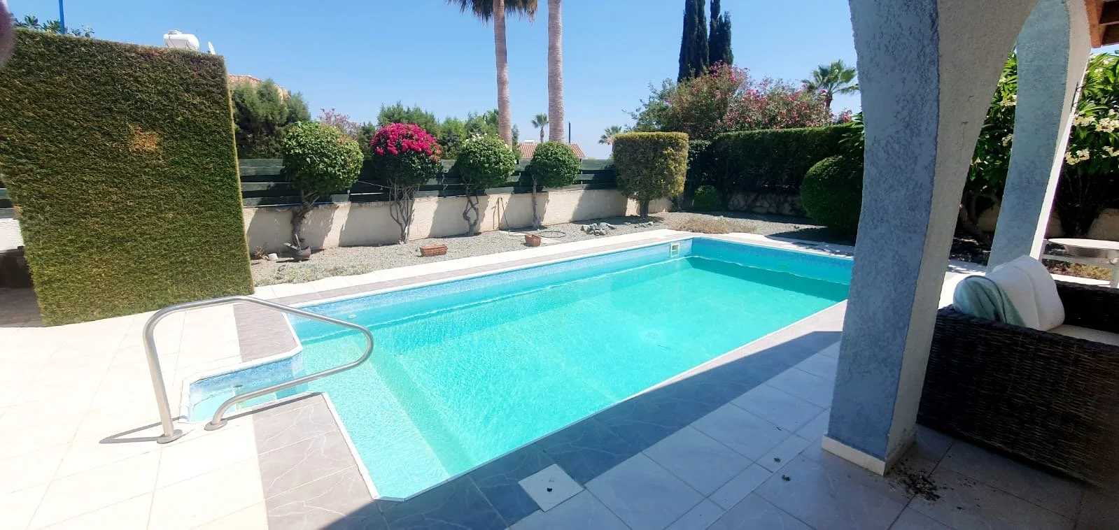 Swimming pool in a backyard surrounded by green bushes and trees, with a seating area on the right.
