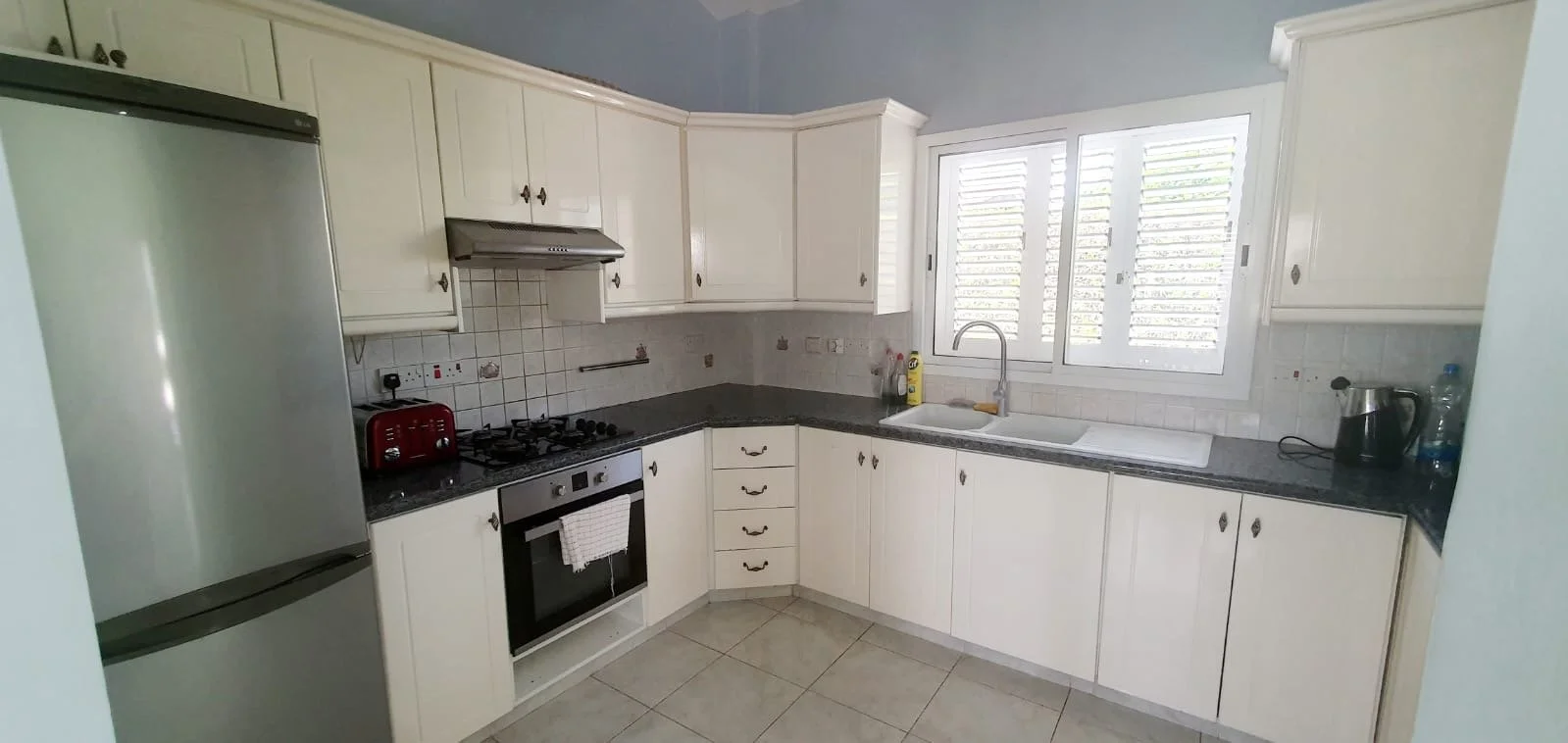 A kitchen with white cabinets, a gray countertop, a silver refrigerator, a red toaster, a stove with an oven, a sink under a window with white shutters, and various kitchen appliances on the counter.