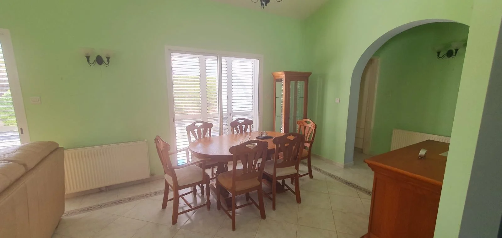 Dining room with green walls, a wooden dining table with six chairs, a glass-front cabinet, a beige sofa partially visible, and a tile floor with decorative border.