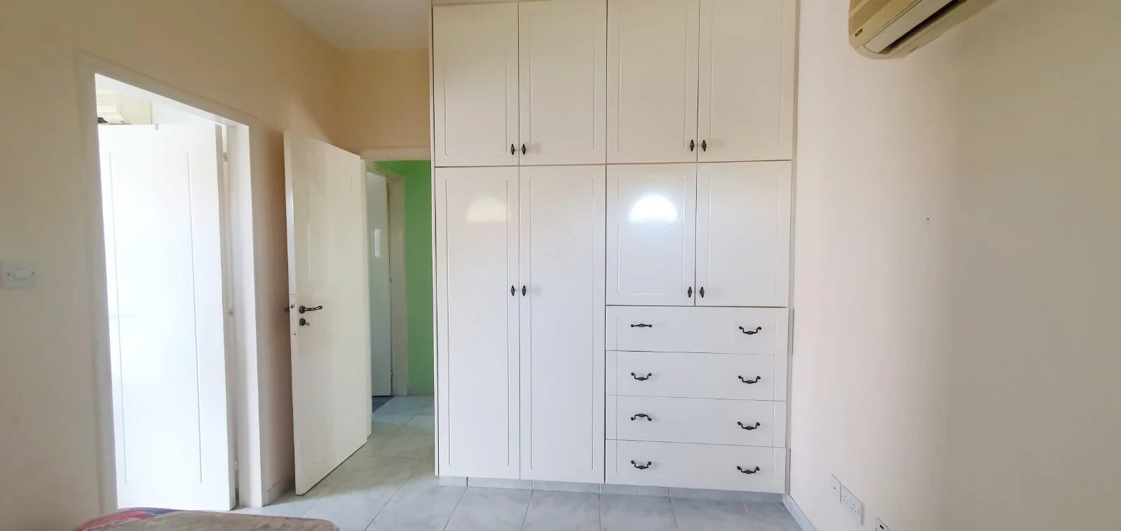 A room with a built-in white cabinet featuring multiple doors and drawers, an air conditioning unit mounted on the wall, and a doorway leading to another room with a green wall.
