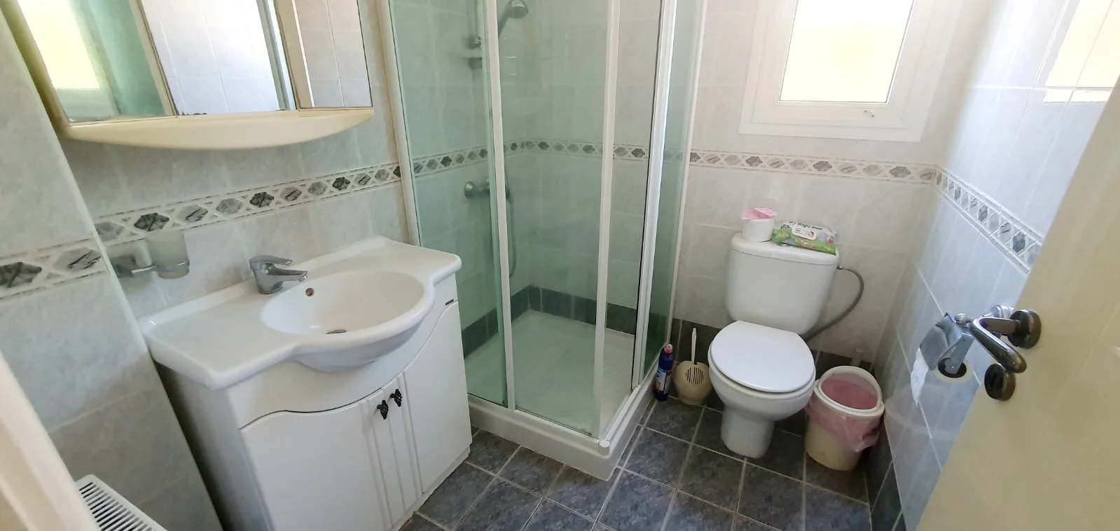 Small bathroom with a shower enclosure, white sink with cabinet, toilet, and window, decorated with beige tiles and a decorative tile border.