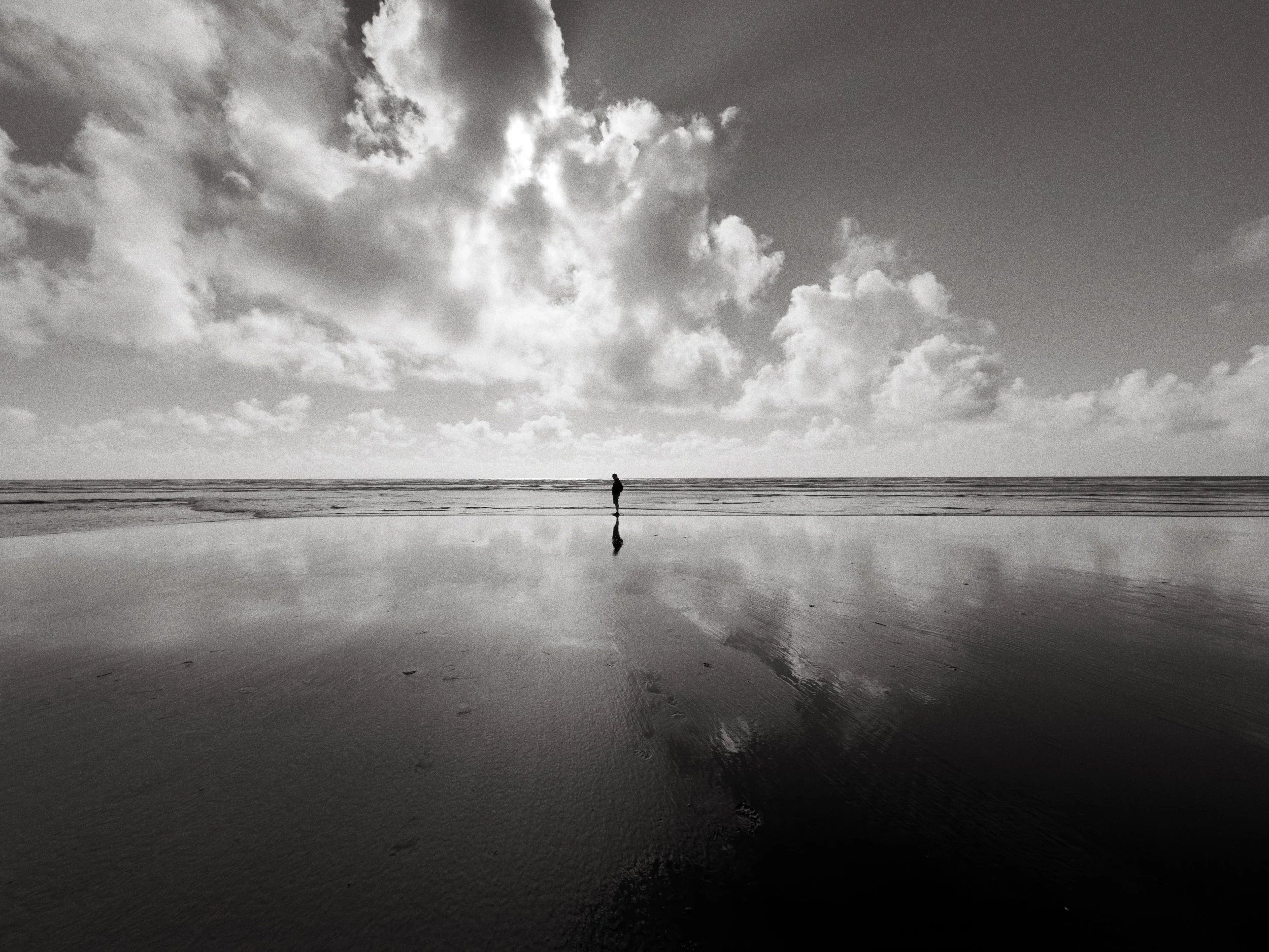 A person walking alone on a beach with a cloudy sky above, in black and white.