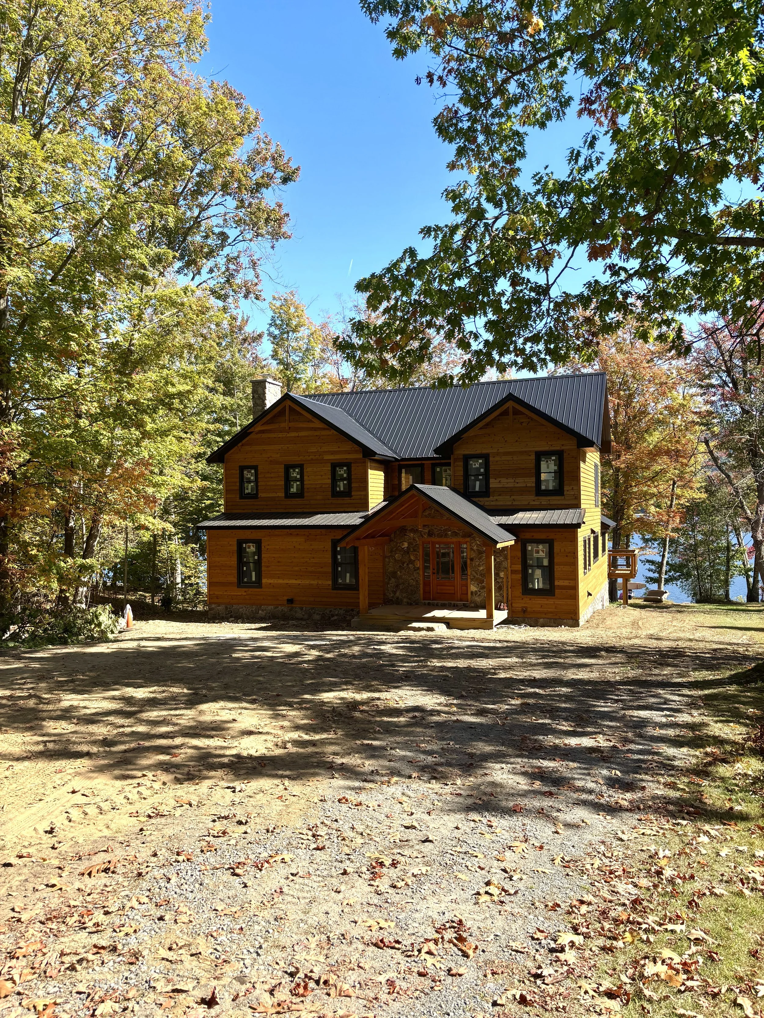 A two-story wooden house with a dark metal roof surrounded by trees with autumn leaves, a clear blue sky, and a gravel driveway in the foreground.