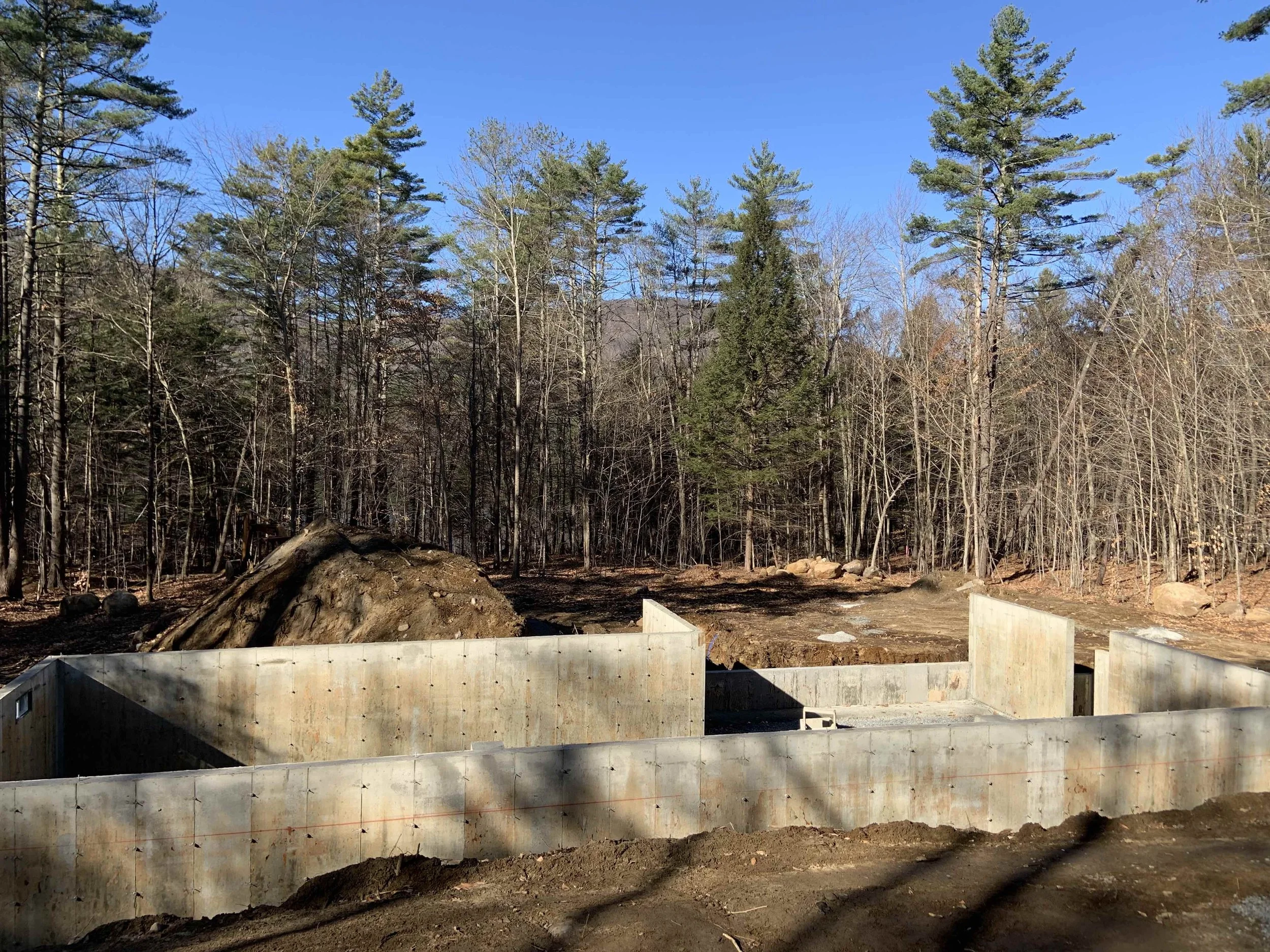 Poured concrete foundation walls on a wooded lot, with cleared and graded land, excavated fill piles, and exposed rock visible around the site.