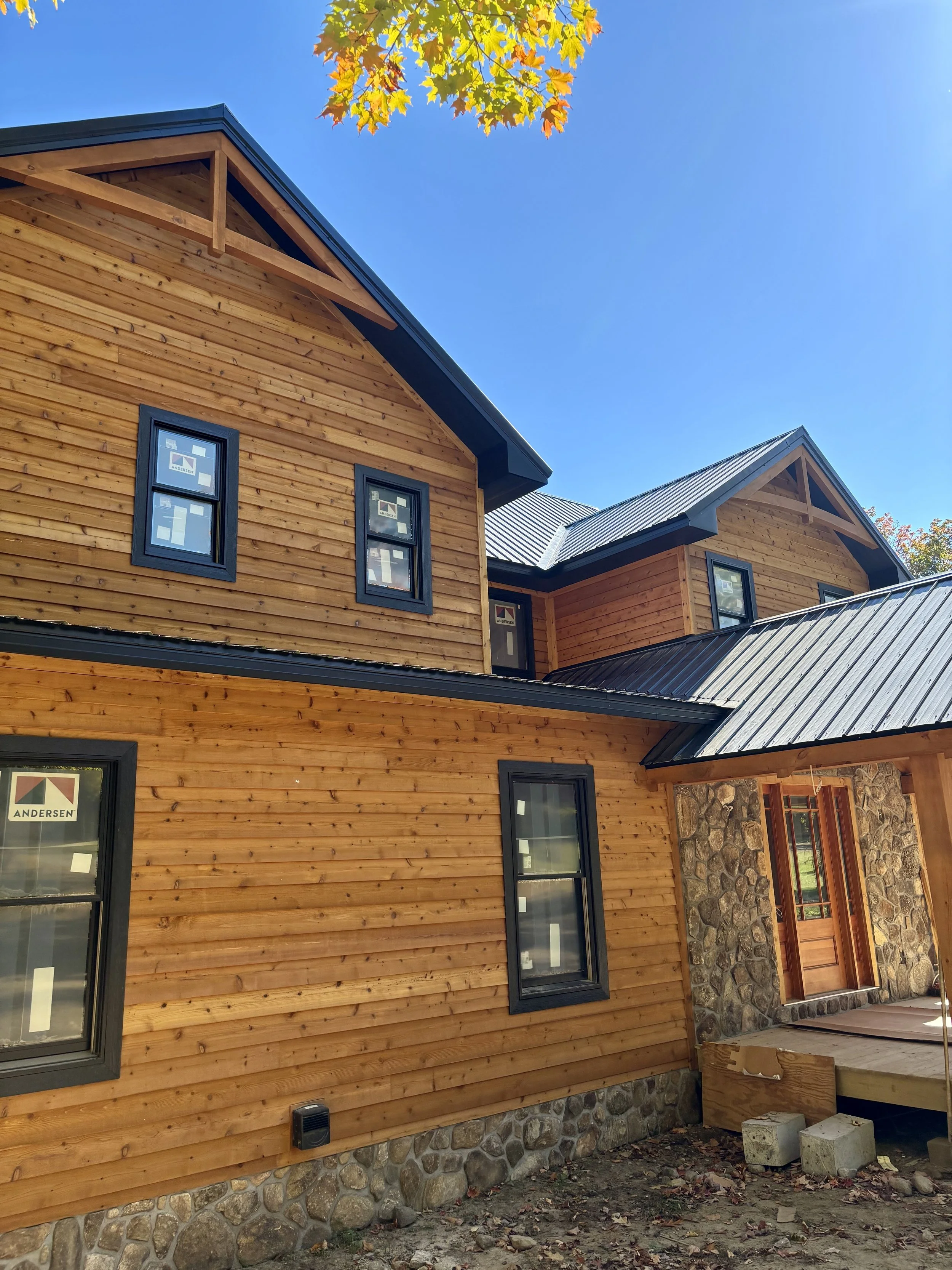 Exterior of a timber-frame home with natural cedar siding, a standing-seam metal roof, stone veneer accents, and an exposed timber gable arch above the entry, still under construction with window protective film in place.