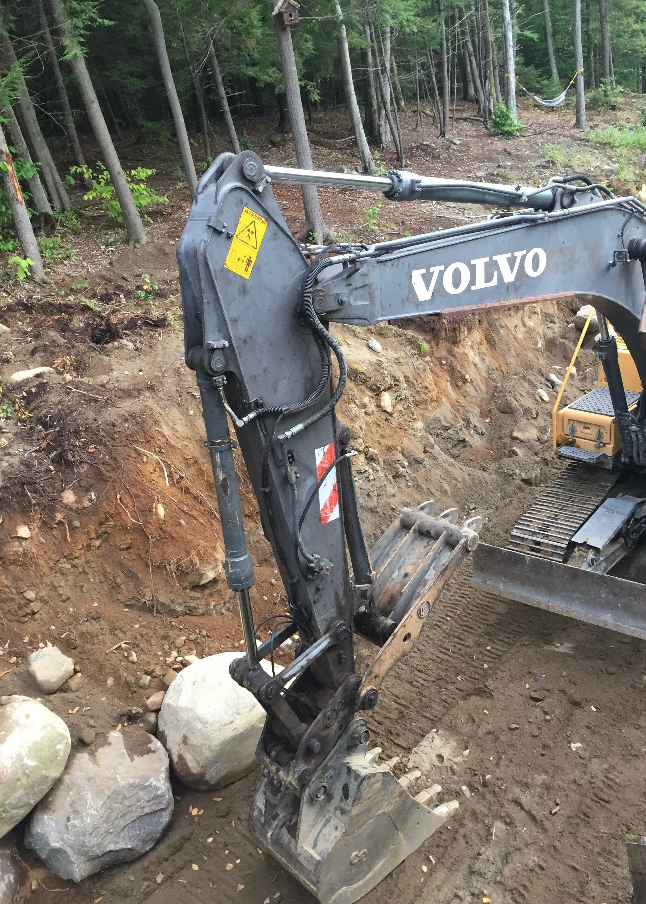 Excavator arm and bucket working a cut bank of rocky soil on a wooded building lot, with large boulders pulled to the side