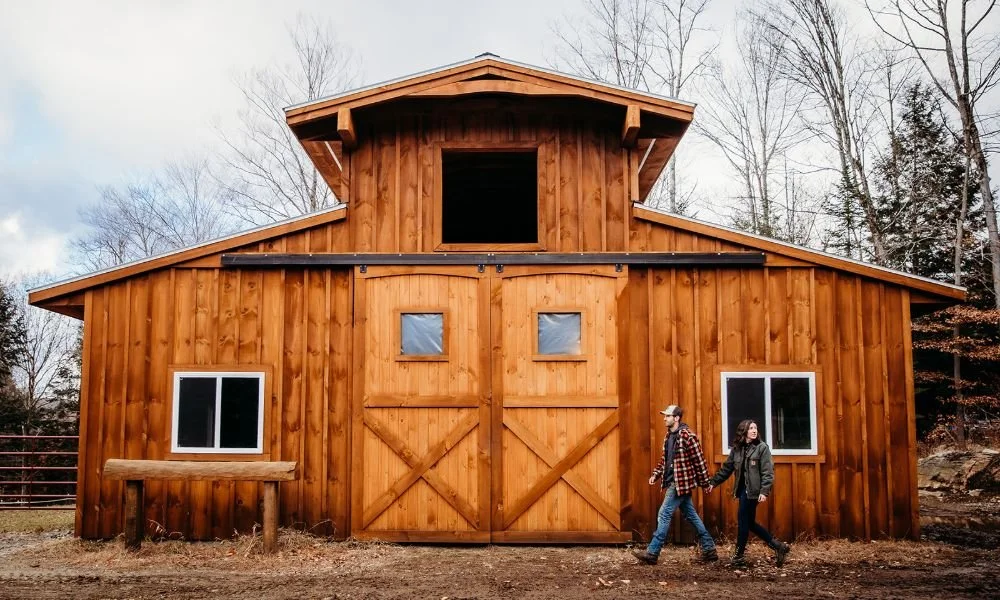 A couple walking in front of a wooden barn with sliding doors, small windows, and a second-story opening, surrounded by trees.
