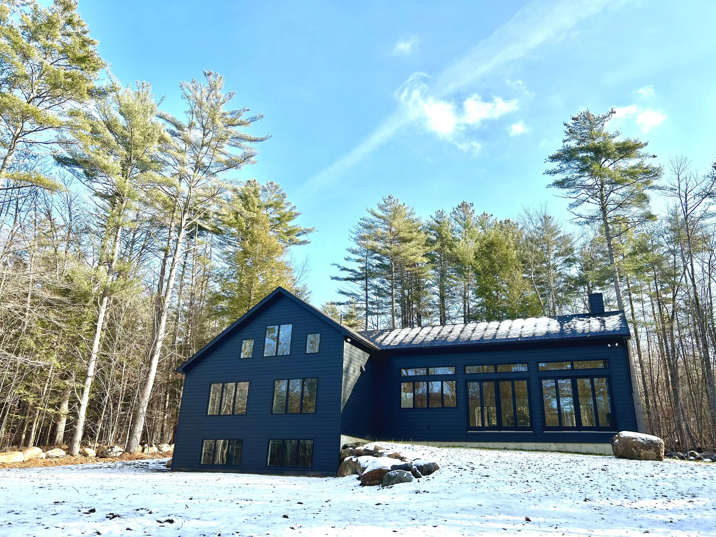 Finished custom home with black siding, black-framed windows, and a snow-dusted metal roof, set among tall pines with a light snow covering the ground.