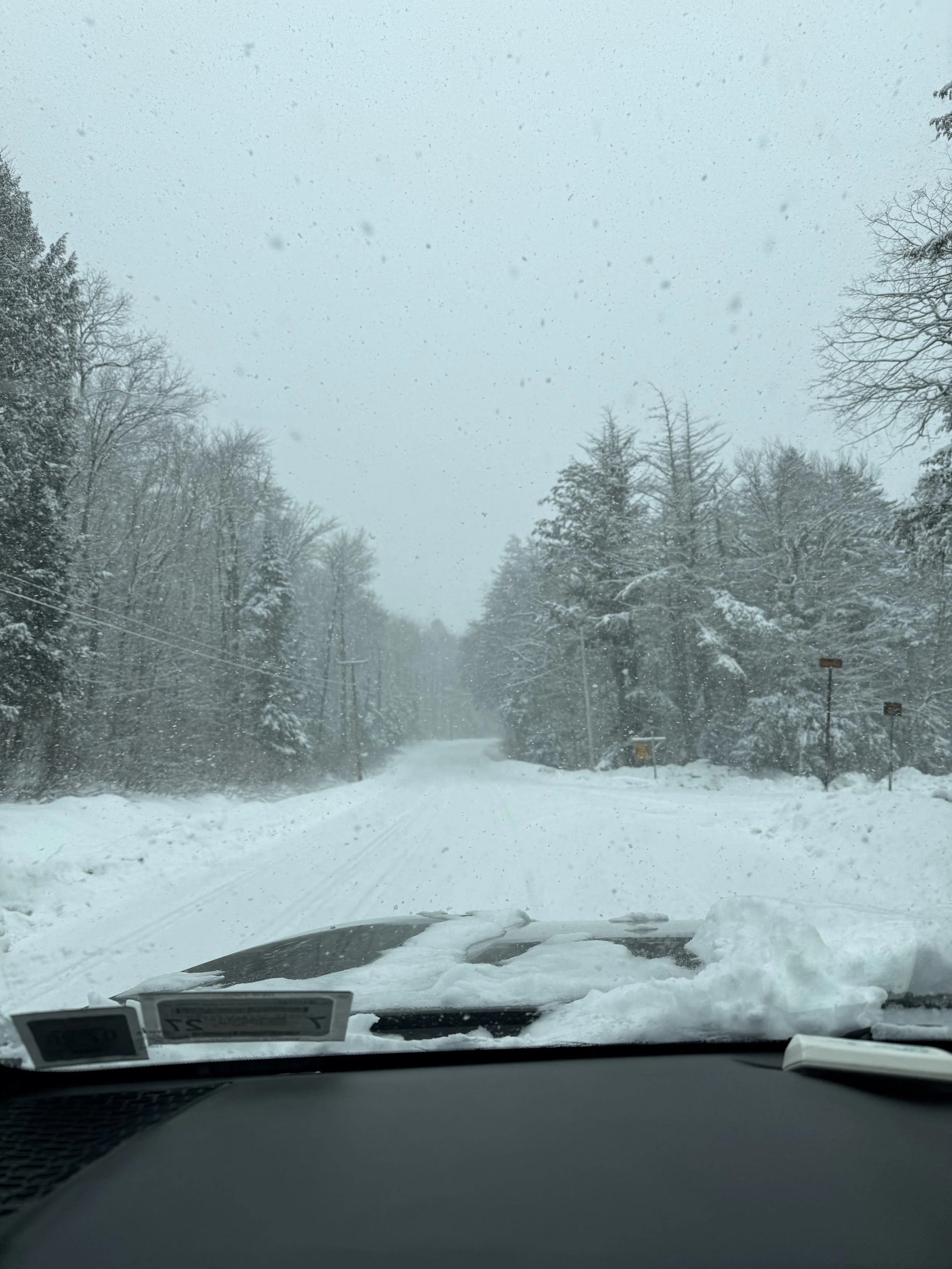 Snow-covered rural road in the southern Adirondacks during a winter storm, viewed from inside a vehicle with snow piled on the hood