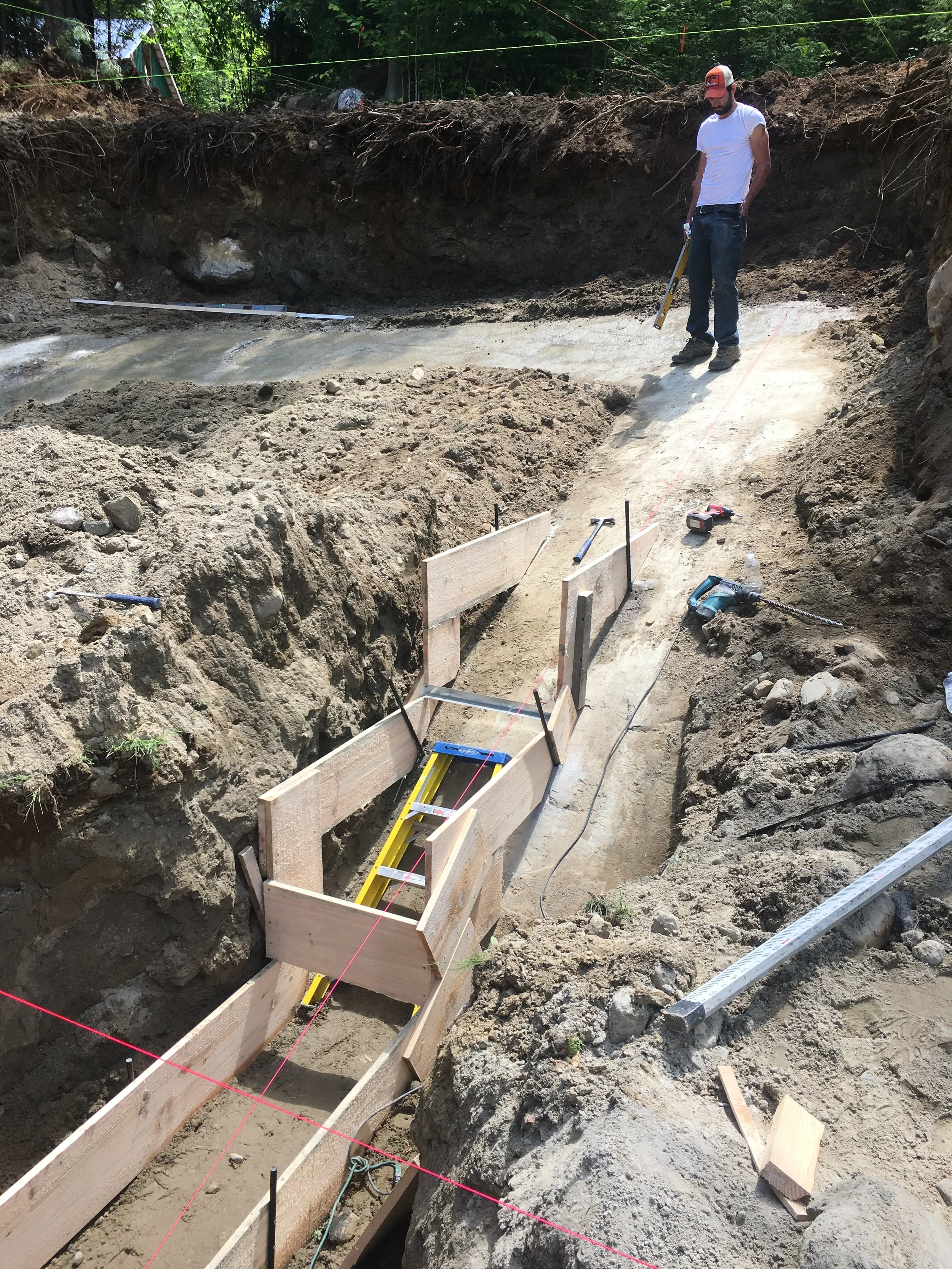 Excavated trench with wood footing forms and rebar stakes set against an exposed ledgerock face, a worker checking grade with a level, and string lines marking the layout for a home addition foundation.