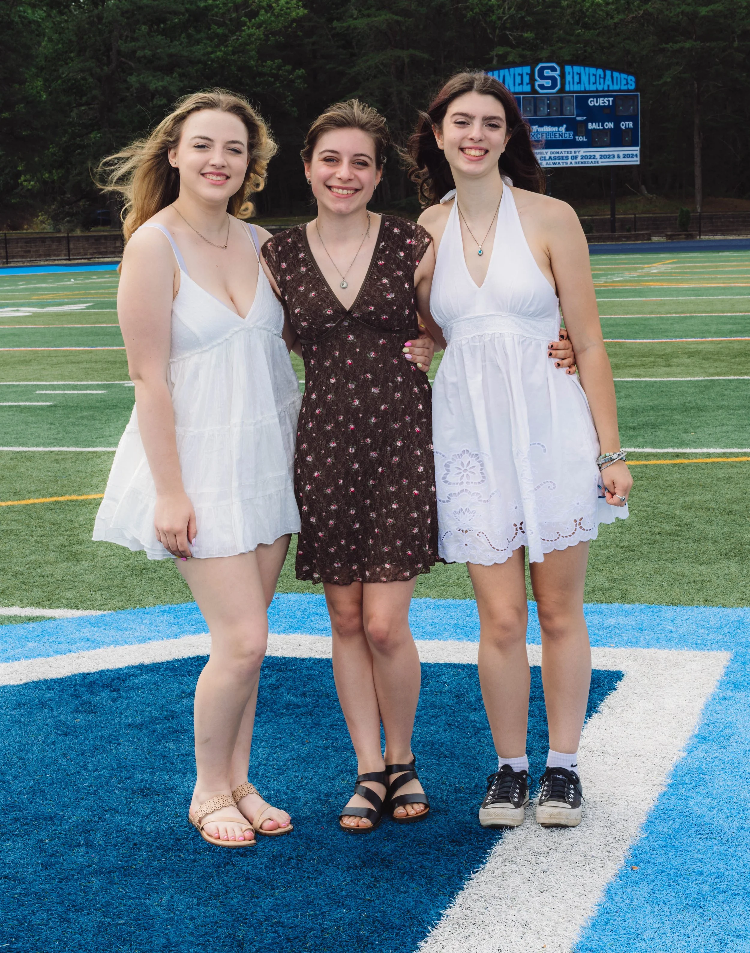 Three young women standing on a football field, smiling, with an electronic scoreboard in the background.