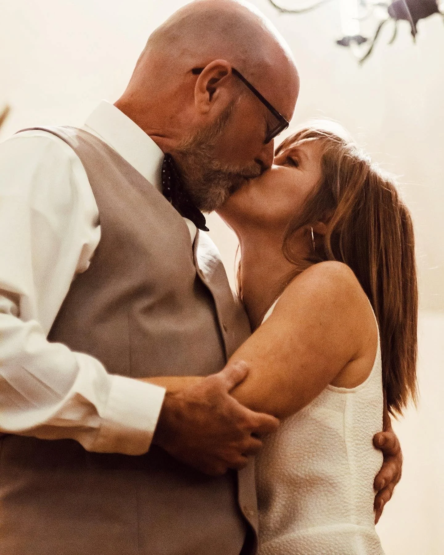 A man with glasses and a beard kissing a woman with brown hair, wearing a white sleeveless dress, in an embrace.