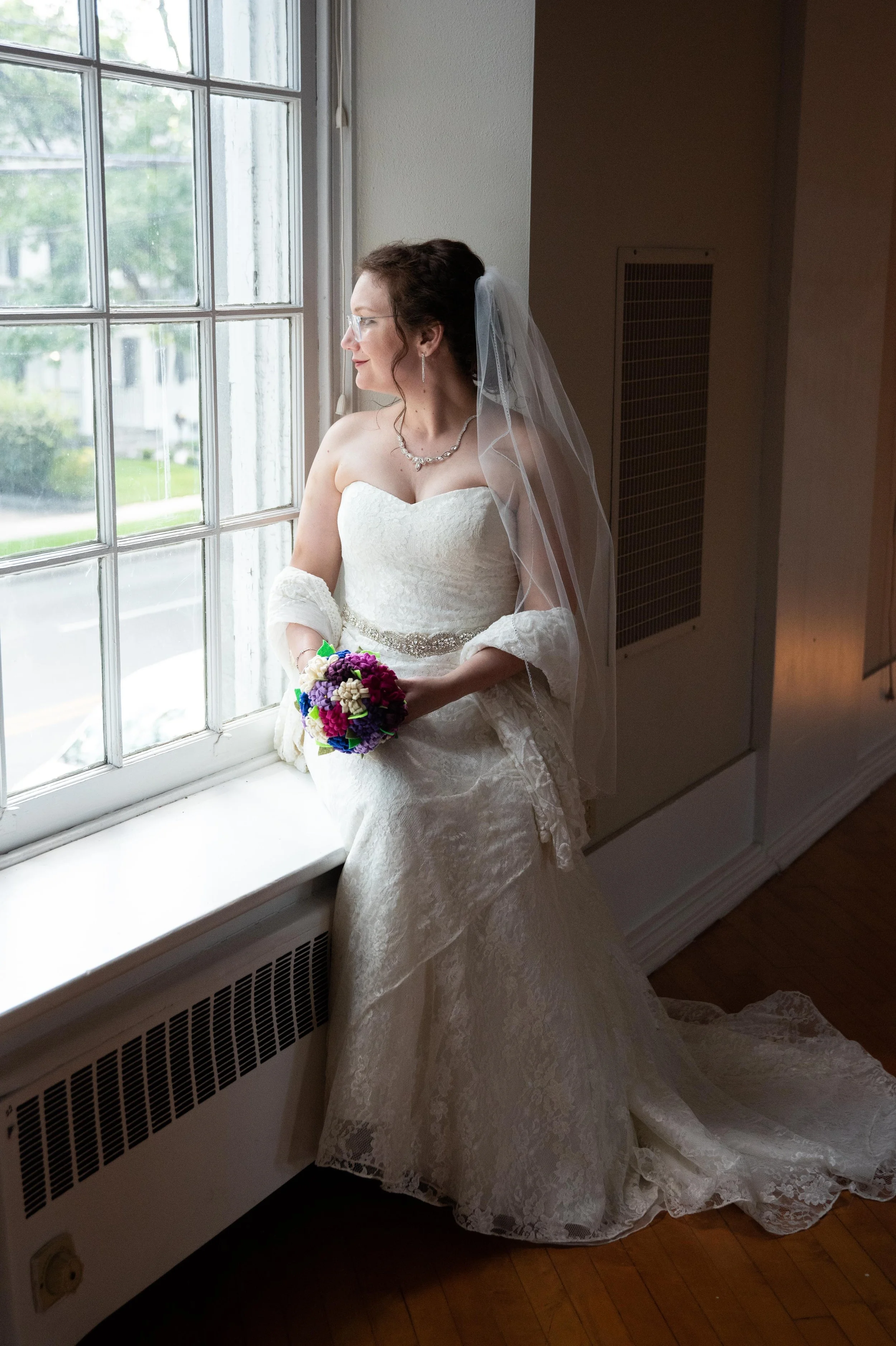 A bride in a white lace wedding dress standing by a window, holding a bouquet, with a veil on her head, looking outside.