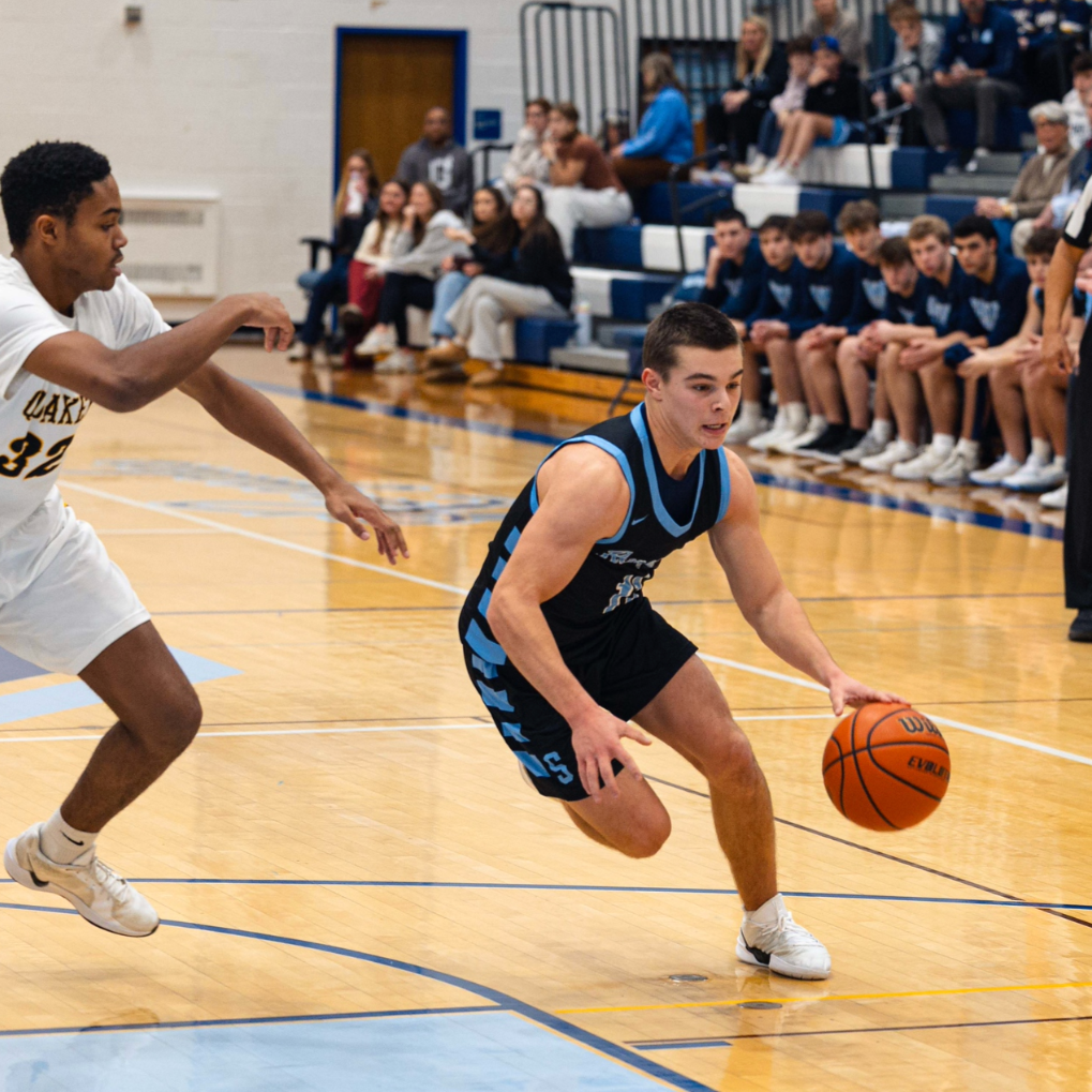 Two high school boys playing basketball during a game inside a gymnasium. One player in a black and blue uniform is dribbling the ball while the other in a white uniform is defending. Spectators sit on bleachers in the background.