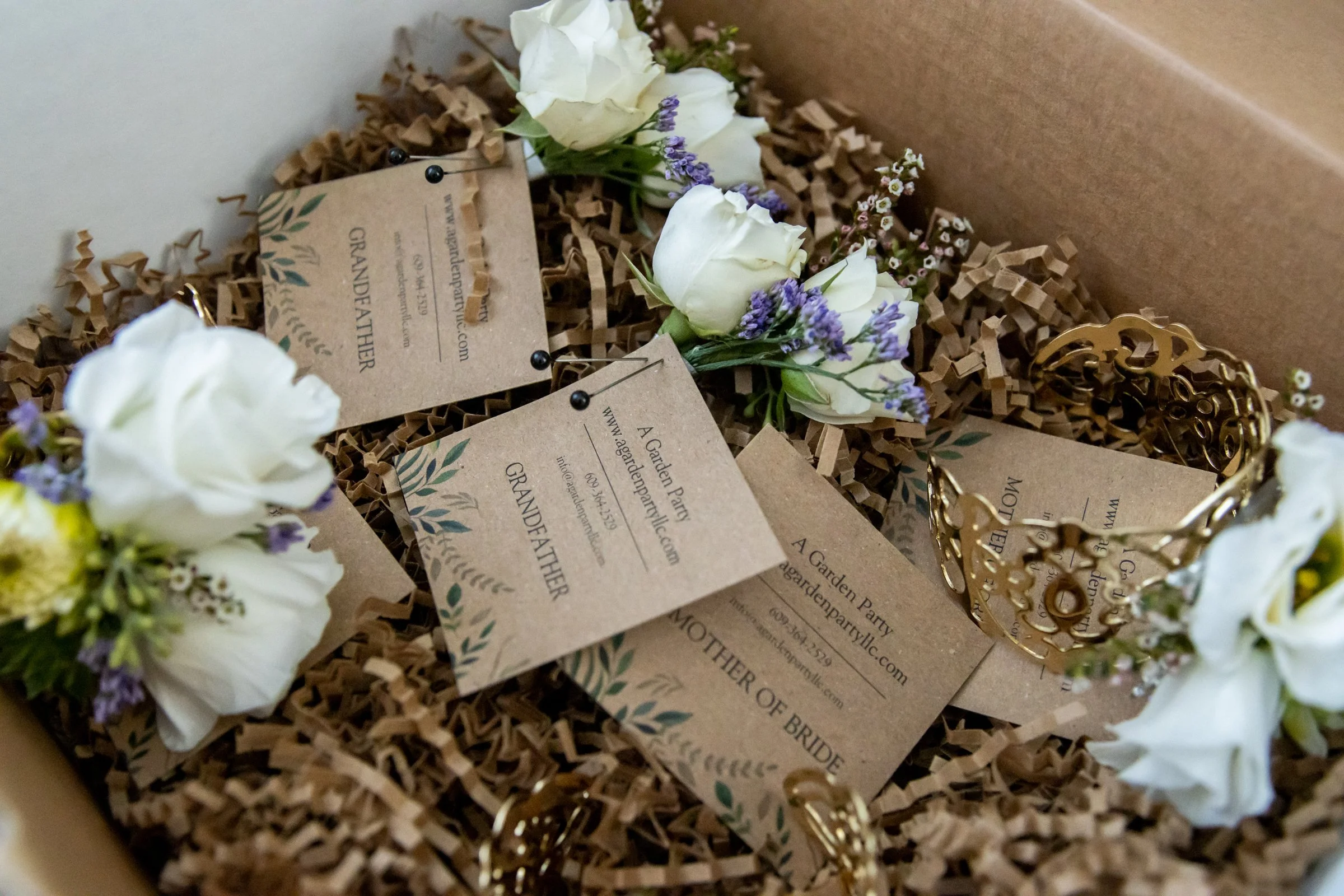 A gift box filled with crinkled brown paper shred and decorated with white roses, purple lavender, and small white flowers, along with cards and a golden decorative piece inside.