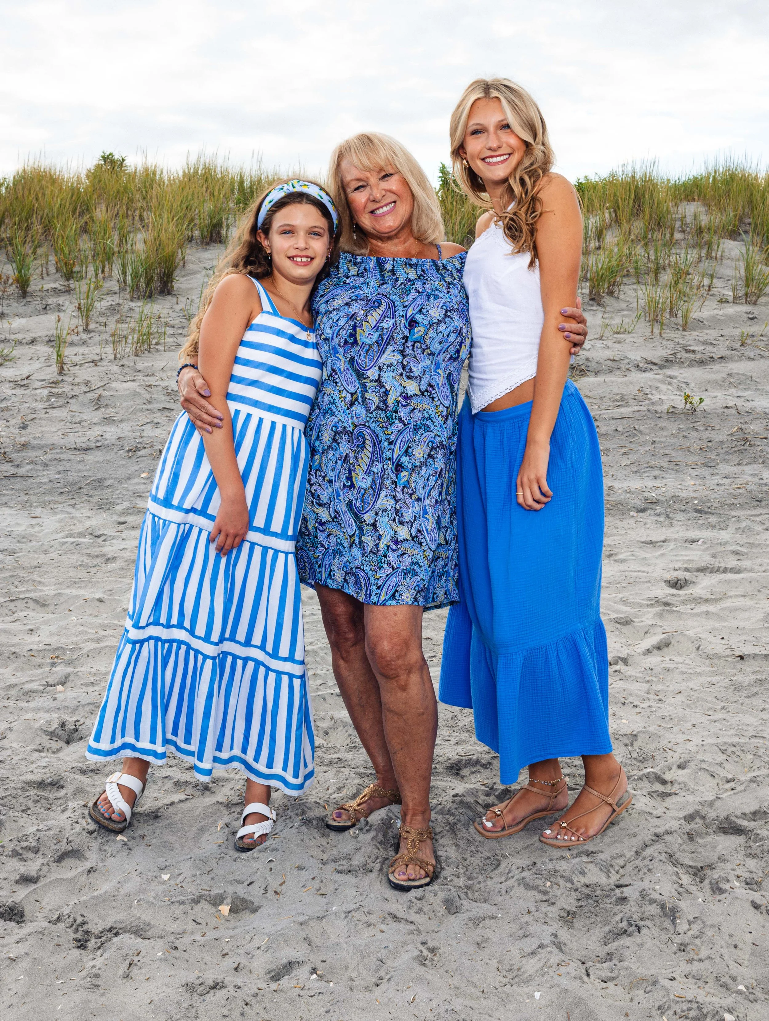 Three women standing on a sandy beach with tall grass in the background, smiling at the camera.