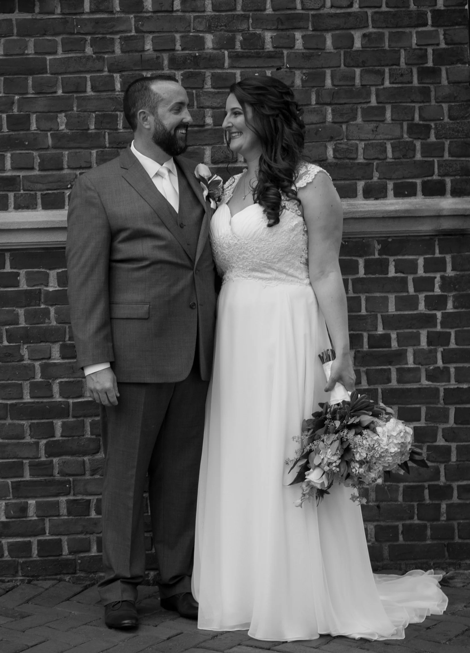 A black and white photograph of a bride and groom standing close in front of a brick wall, smiling at each other. The groom is in a suit with a boutonniere, and the bride is in a wedding dress holding a bouquet of flowers.