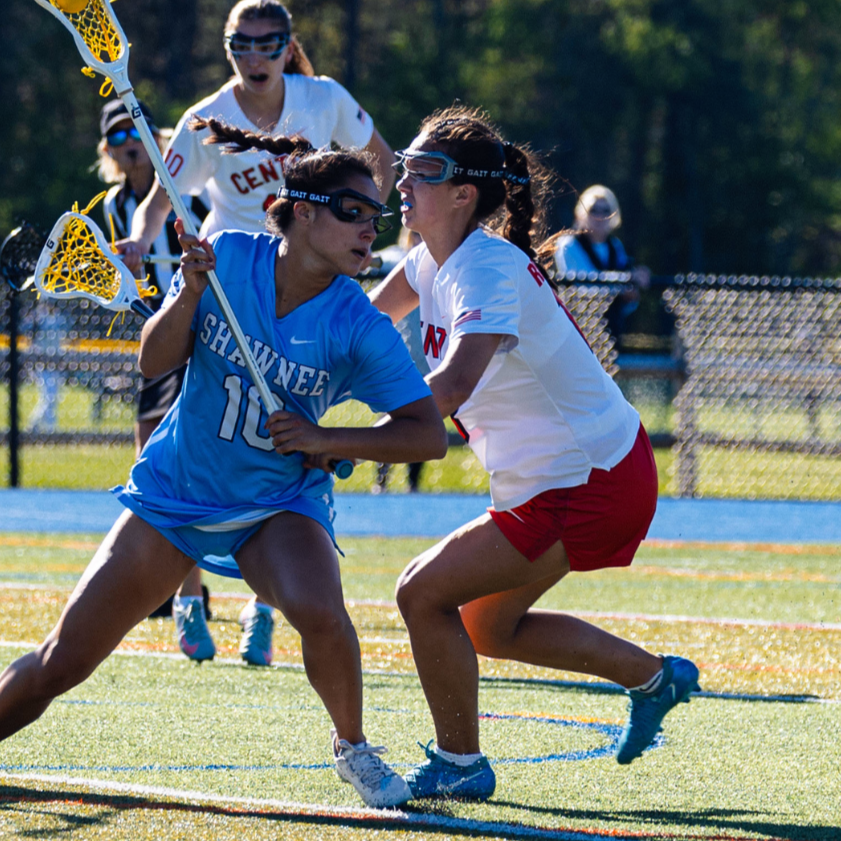 Two female lacrosse players in action during a game, one in a blue jersey with number 10, and the other in a white jersey with red shorts, battling for possession with lacrosse sticks on a sunny field.