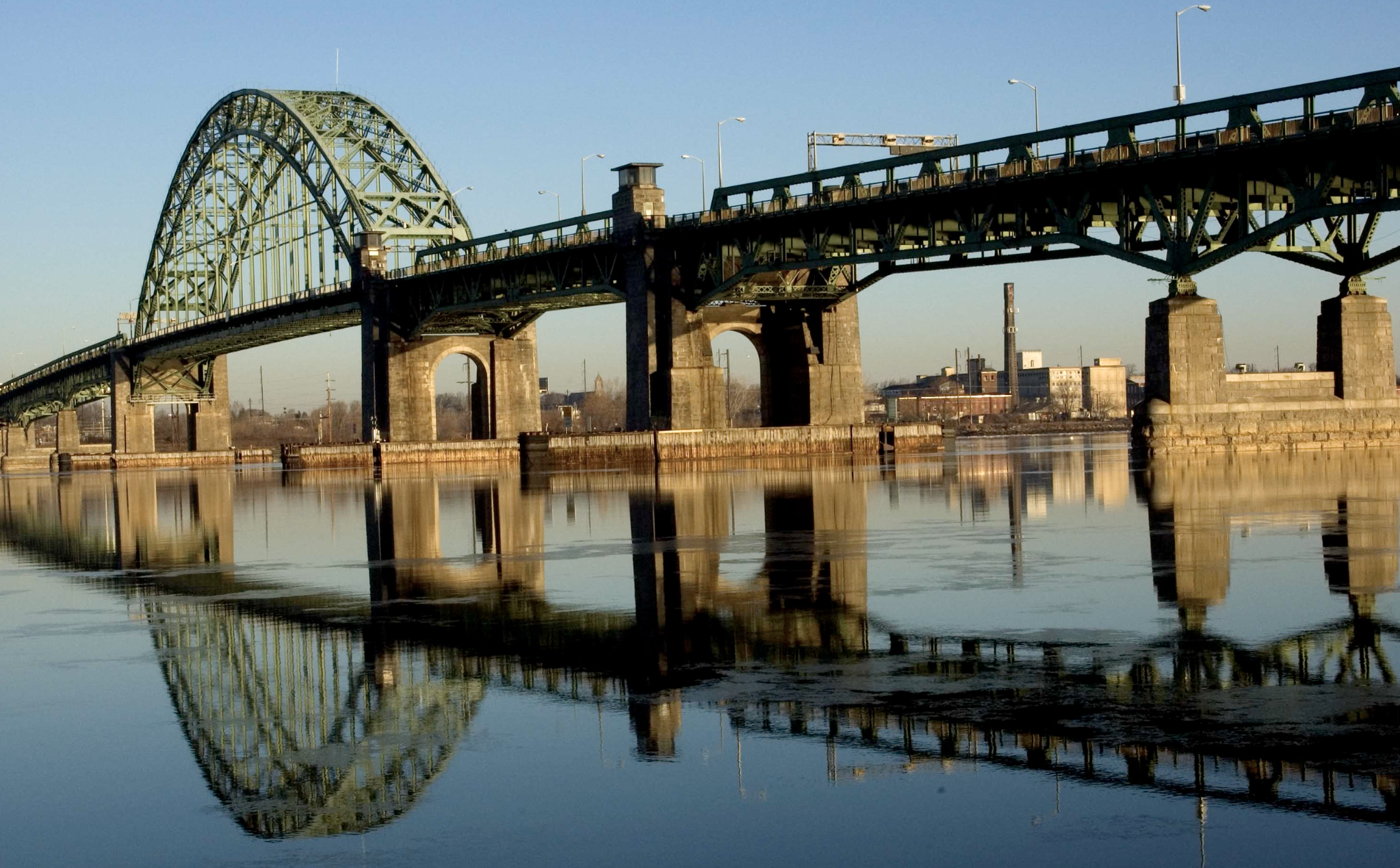 A bridge over a body of water with city buildings in the background, reflecting in the water on a clear day.