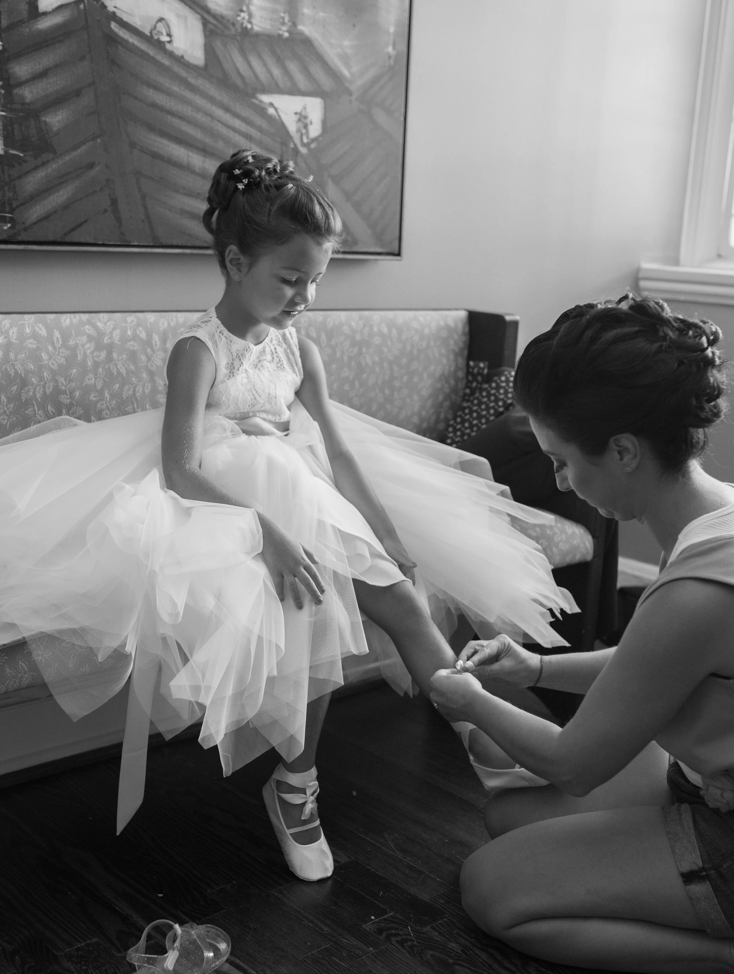 A young girl in a ballet costume getting her foot wrapped by a woman, likely a ballet instructor or parent, while seated on a bench in a room with a framed picture or painting on the wall behind them.