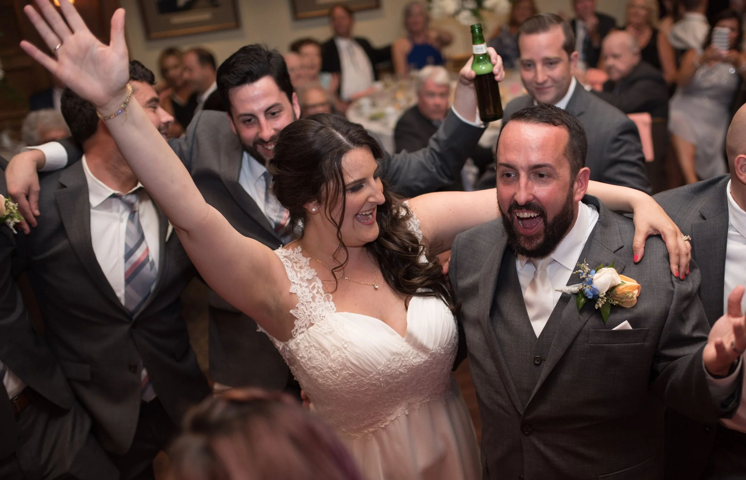 People dancing and celebrating at a wedding reception, including a woman in a white dress and men in suits, some holding drinks, smiling and enjoying the moment.