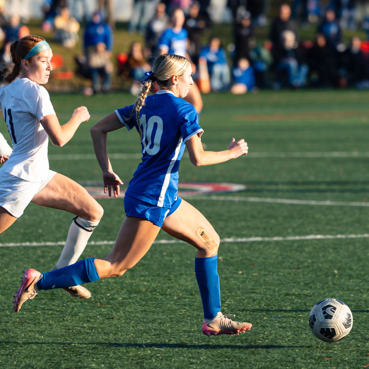Female soccer players chasing the ball during a game on a green field with spectators in the background.