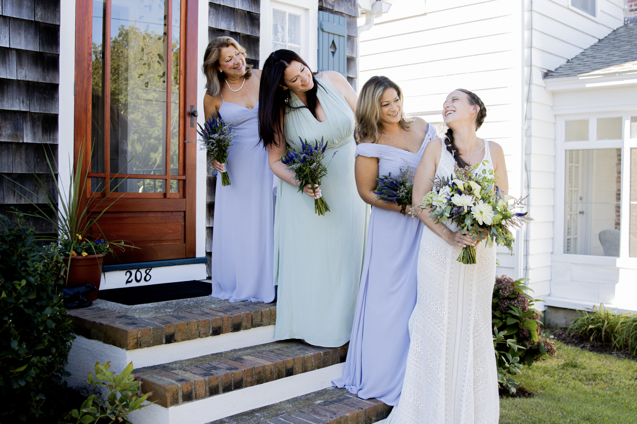 Group of women in pastel dresses holding bouquets, smiling and standing on brick steps outside a house.