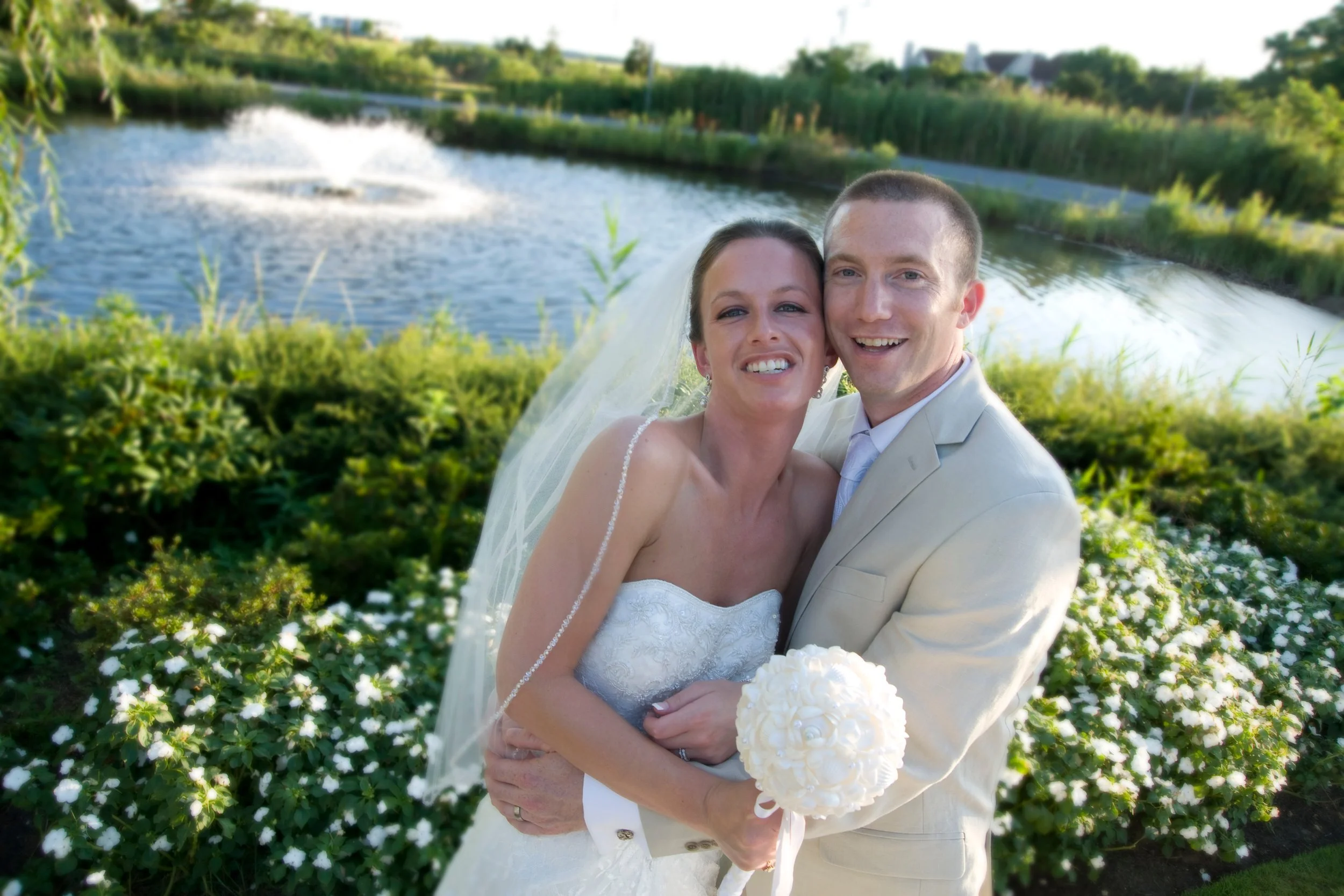 A newlywed couple in wedding attire posing outdoors by a pond with greenery and fountain in the background, smiling at the camera.