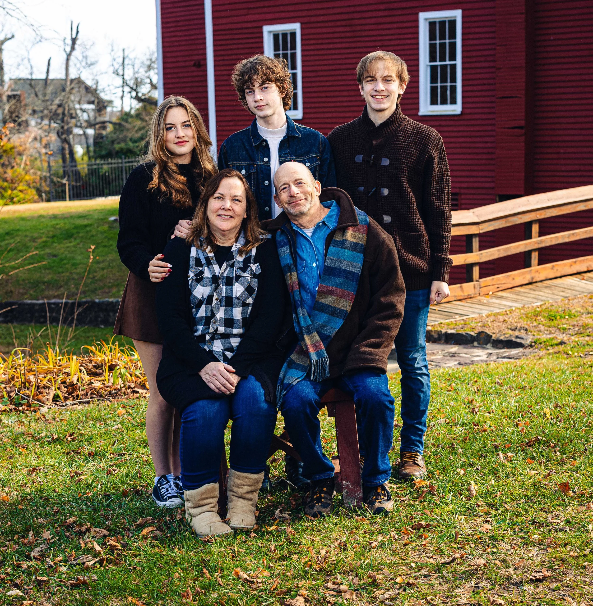 Family portrait outdoors in autumn with four members standing behind a seated older couple, in front of a red house with white trim, surrounded by grass, fallen leaves, and trees.