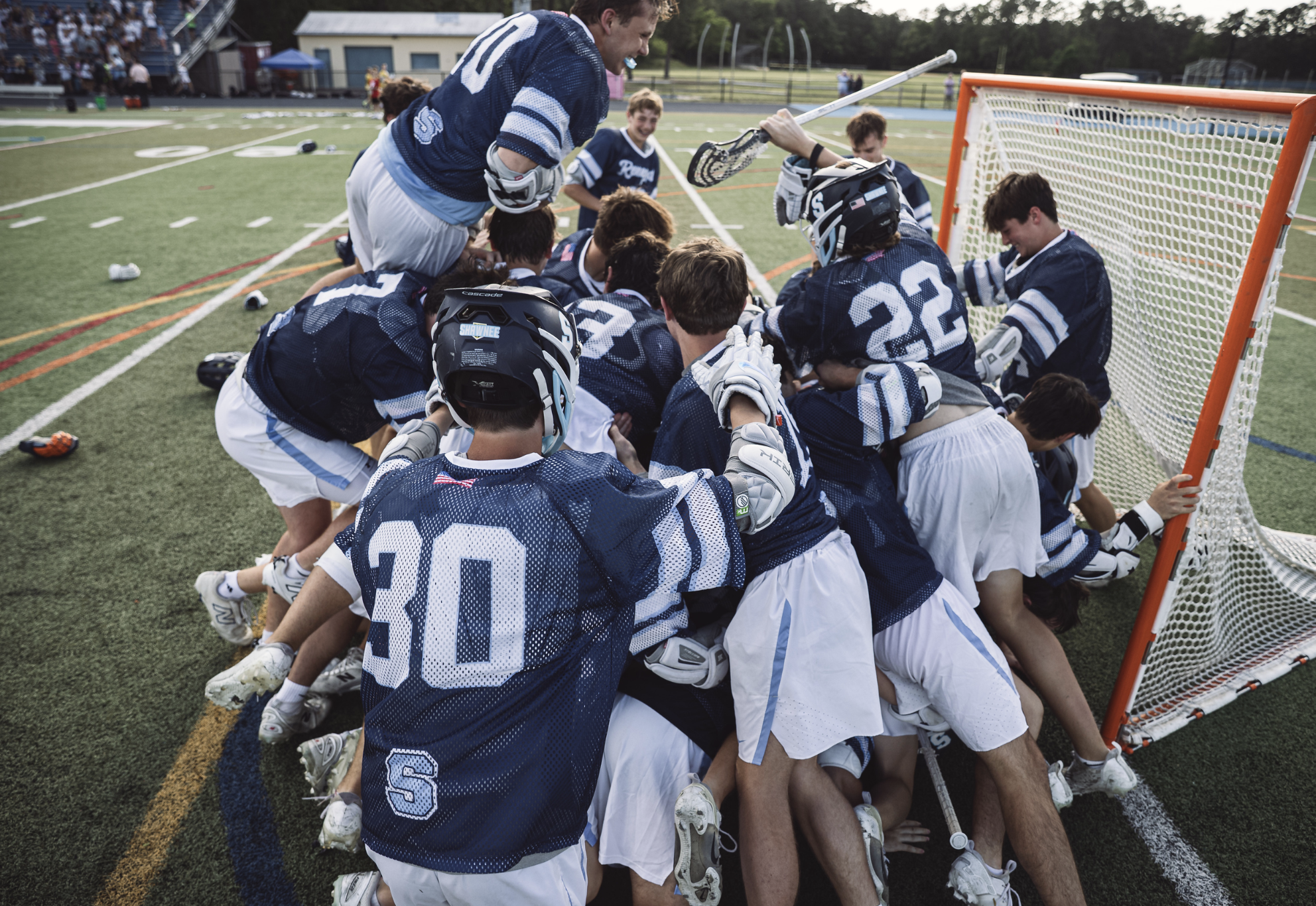 High school boys' lacrosse team celebrating together on a sports field after a game, hugging and high-fiving near the goalpost.