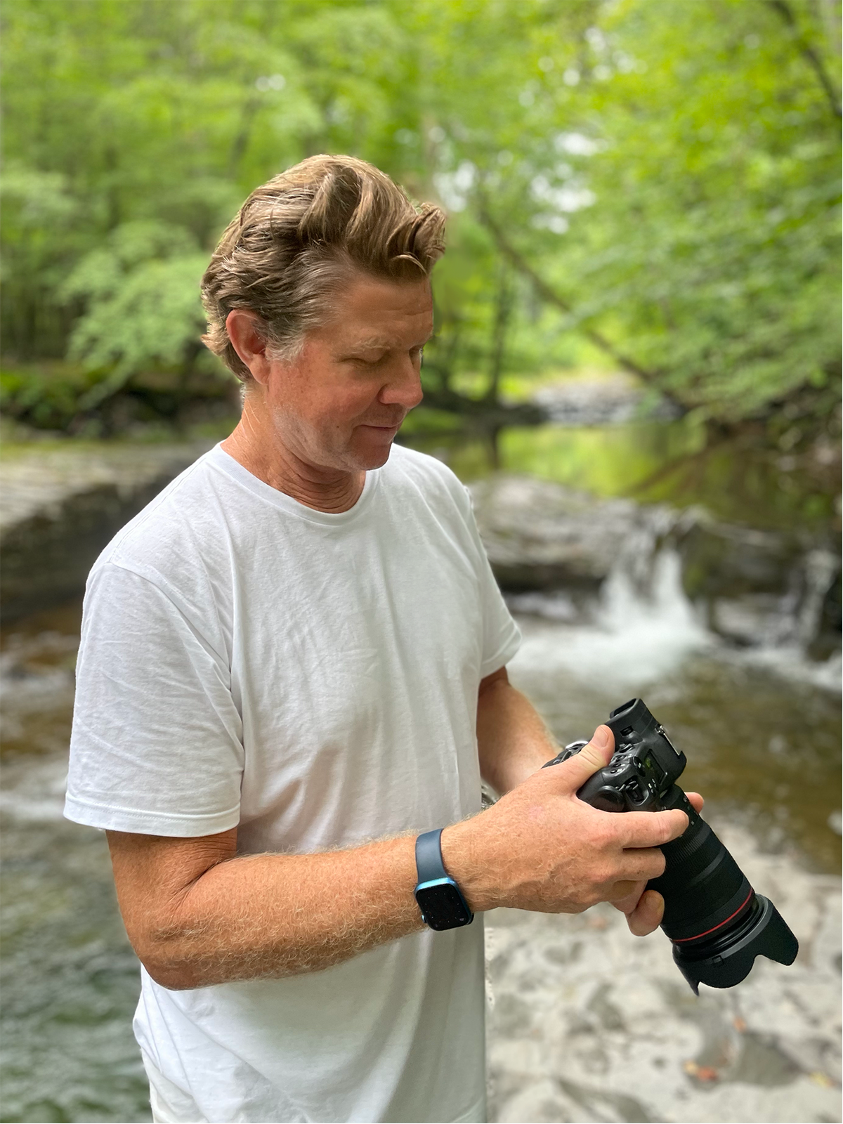 A man with wavy brown hair in a white t-shirt holding a professional camera in a forested area near a stream.