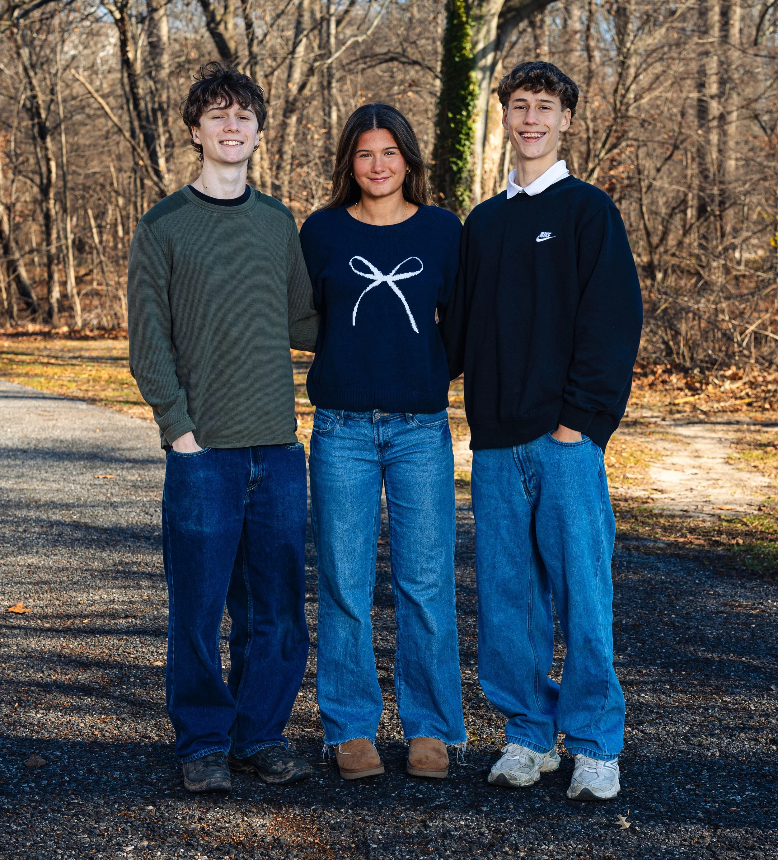 Three teenagers standing outdoors in autumn, smiling and looking at the camera, in a wooded park with bare trees.