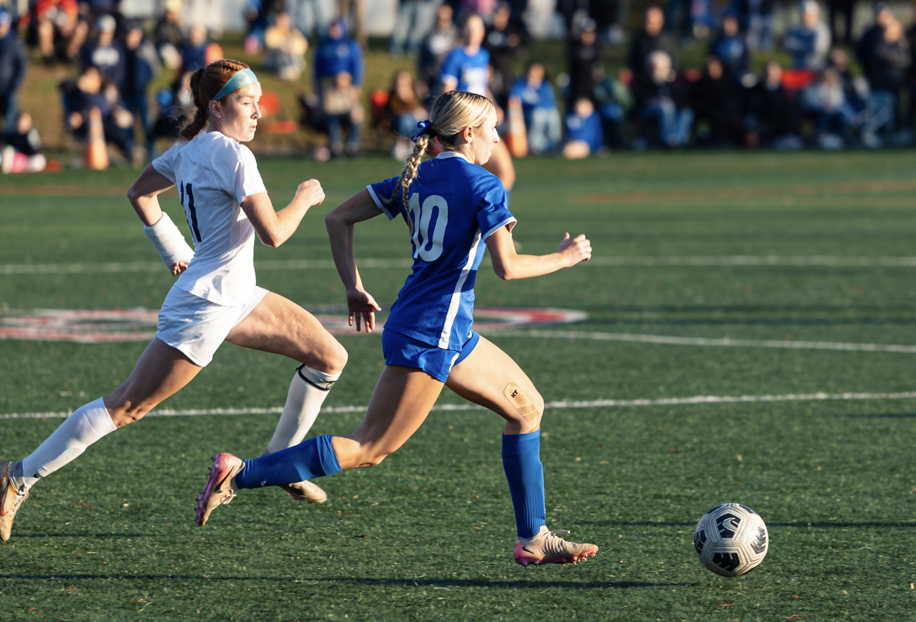 Women playing soccer on a field with spectators in the background.