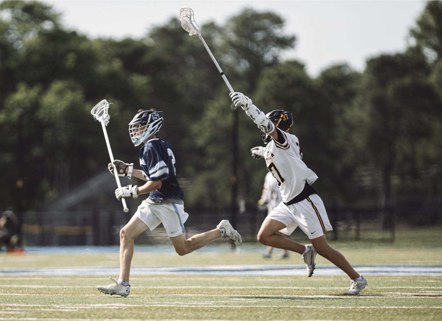 Two lacrosse players in action on the field, with one player in a blue jersey and another in a white jersey, both wearing helmets and holding lacrosse sticks.