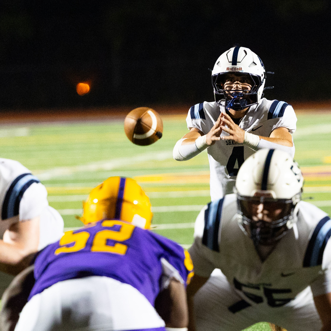 A football player in a white and navy uniform prepares to catch a football during a night game, with opponents in purple and yellow uniforms in front of him.